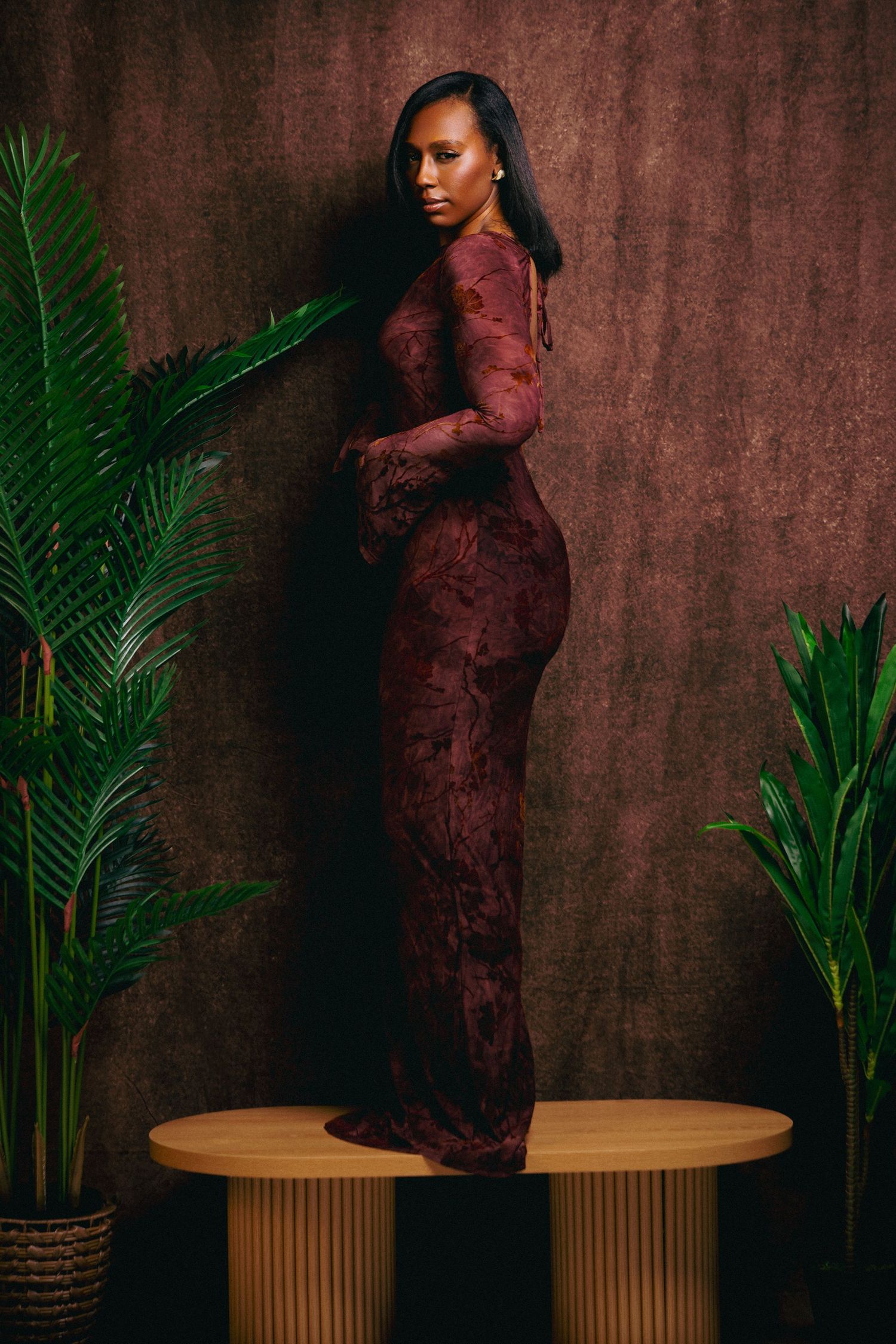 A woman in a fitted burgundy lace gown poses elegantly on a wooden table surrounded by tropical plants.