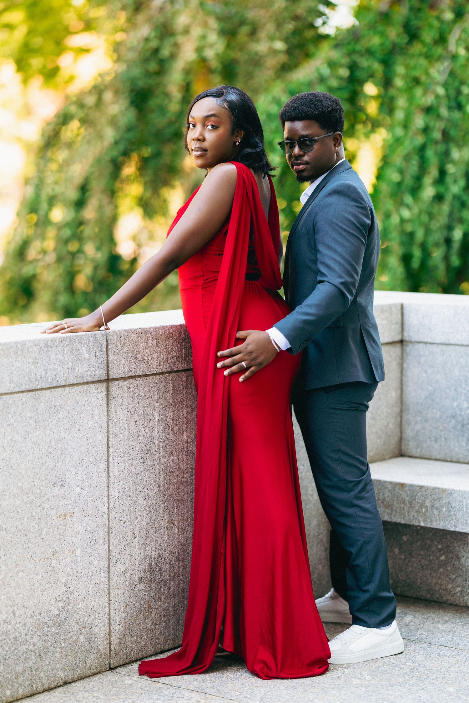 Elegant couple posing outdoors, woman in red gown and man in gray suit with sunglasses, against stone wall.