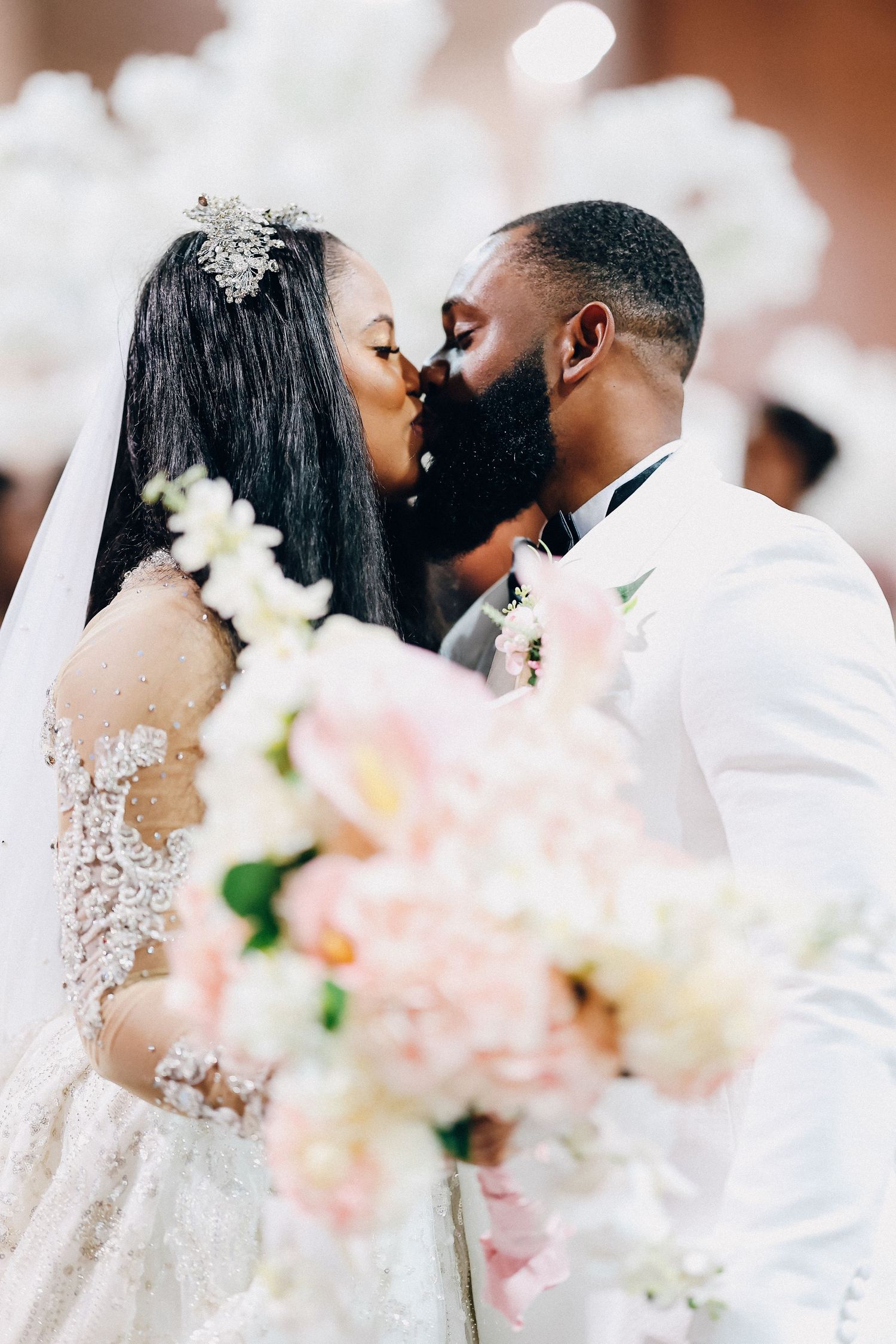 African American bride and groom share first kiss at altar, surrounded by white floral decorations.