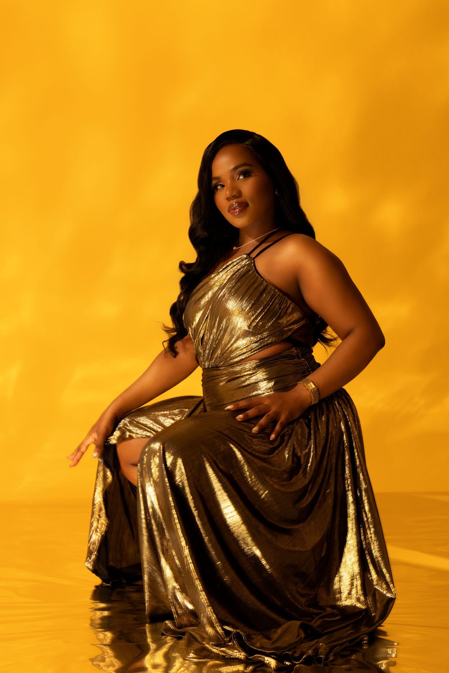 A woman in a gold metallic halter gown poses seated against a vibrant yellow background.