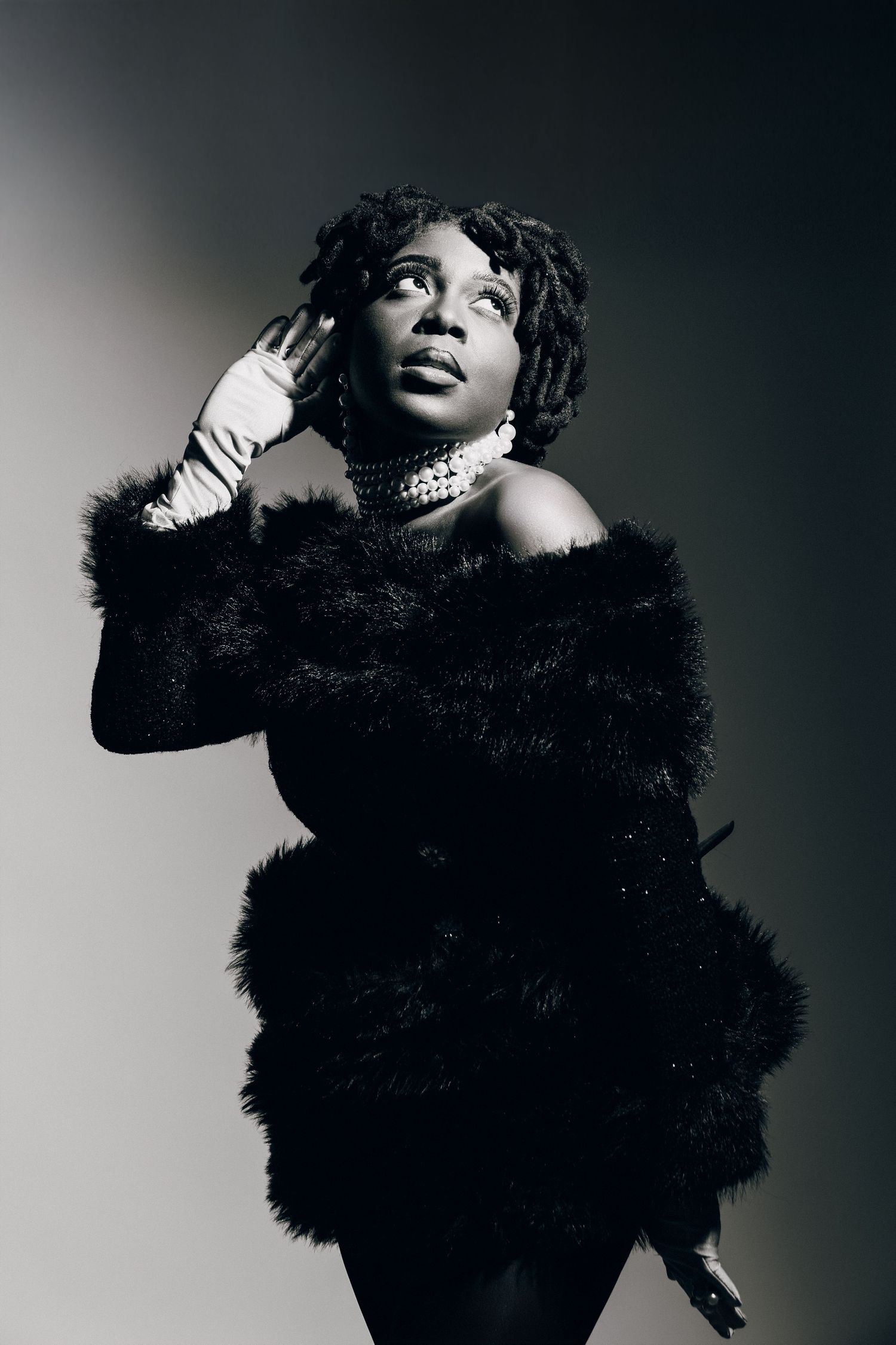 Black and white portrait of a woman in a fur coat and pearl necklace posing elegantly in a studio.