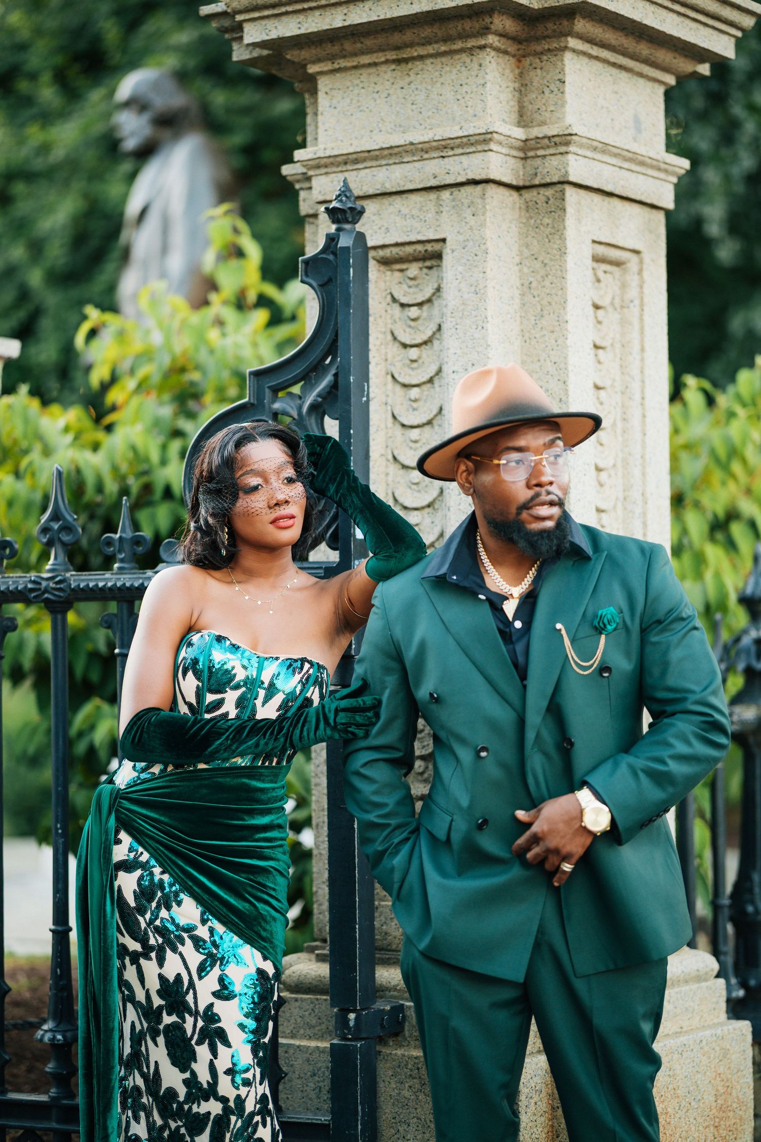Sophisticated Black couple in coordinating emerald green outfits posing by ornate iron gate outdoors.
