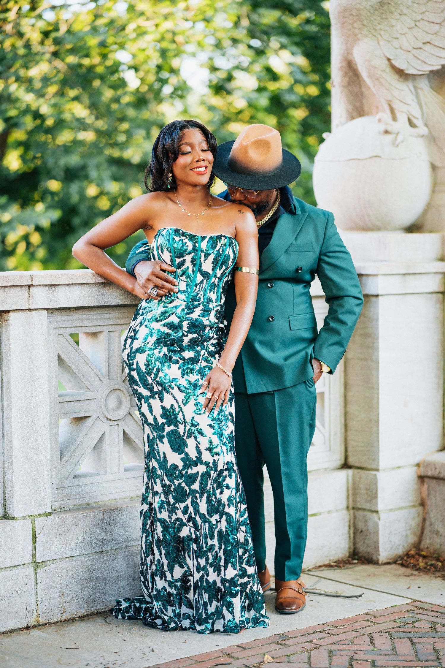 Stylish couple laughing together outdoors, woman in green floral mermaid gown and man in matching teal double-breasted suit and hat.