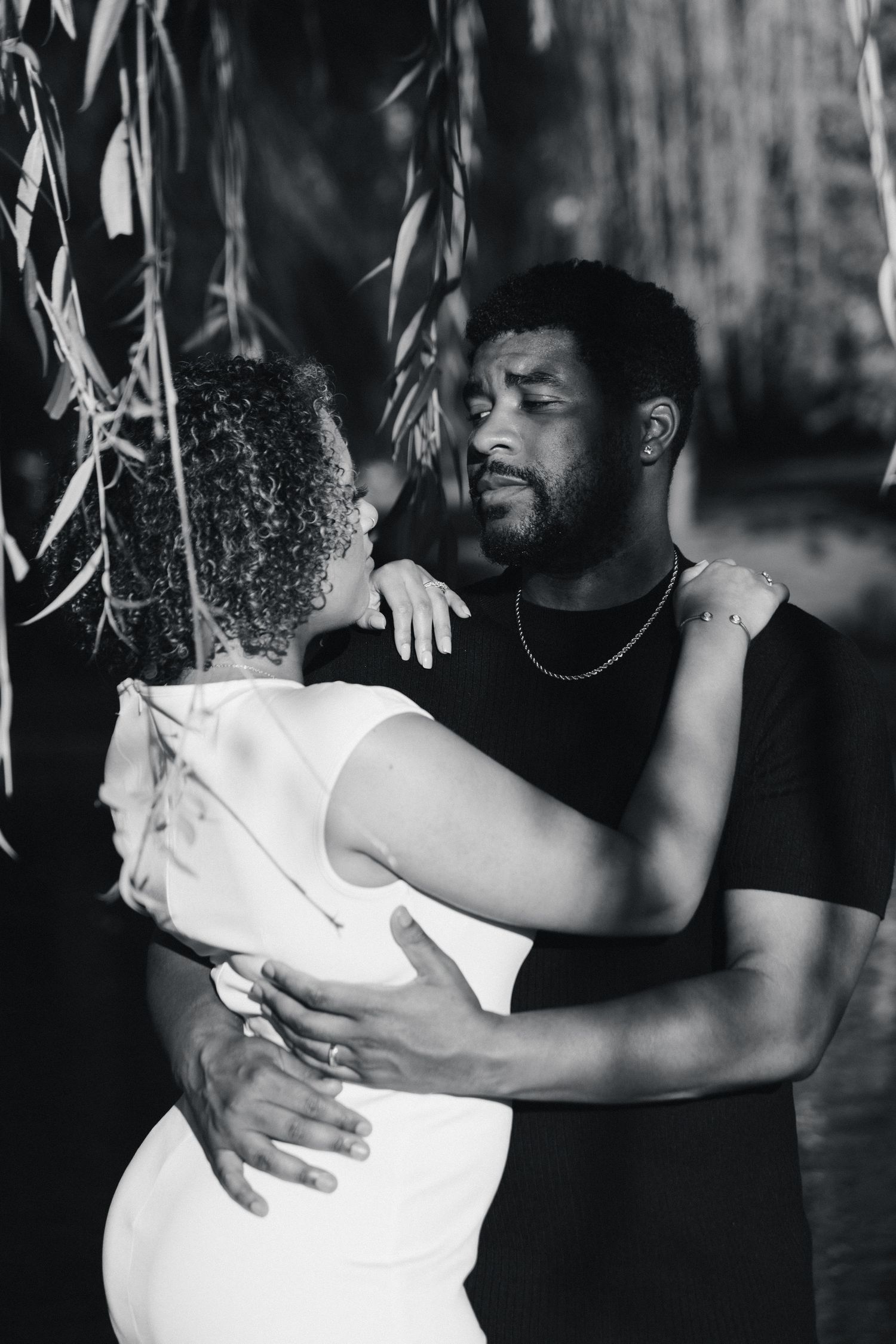 Black and white photo of a couple embracing tenderly under willow tree branches in an intimate moment.