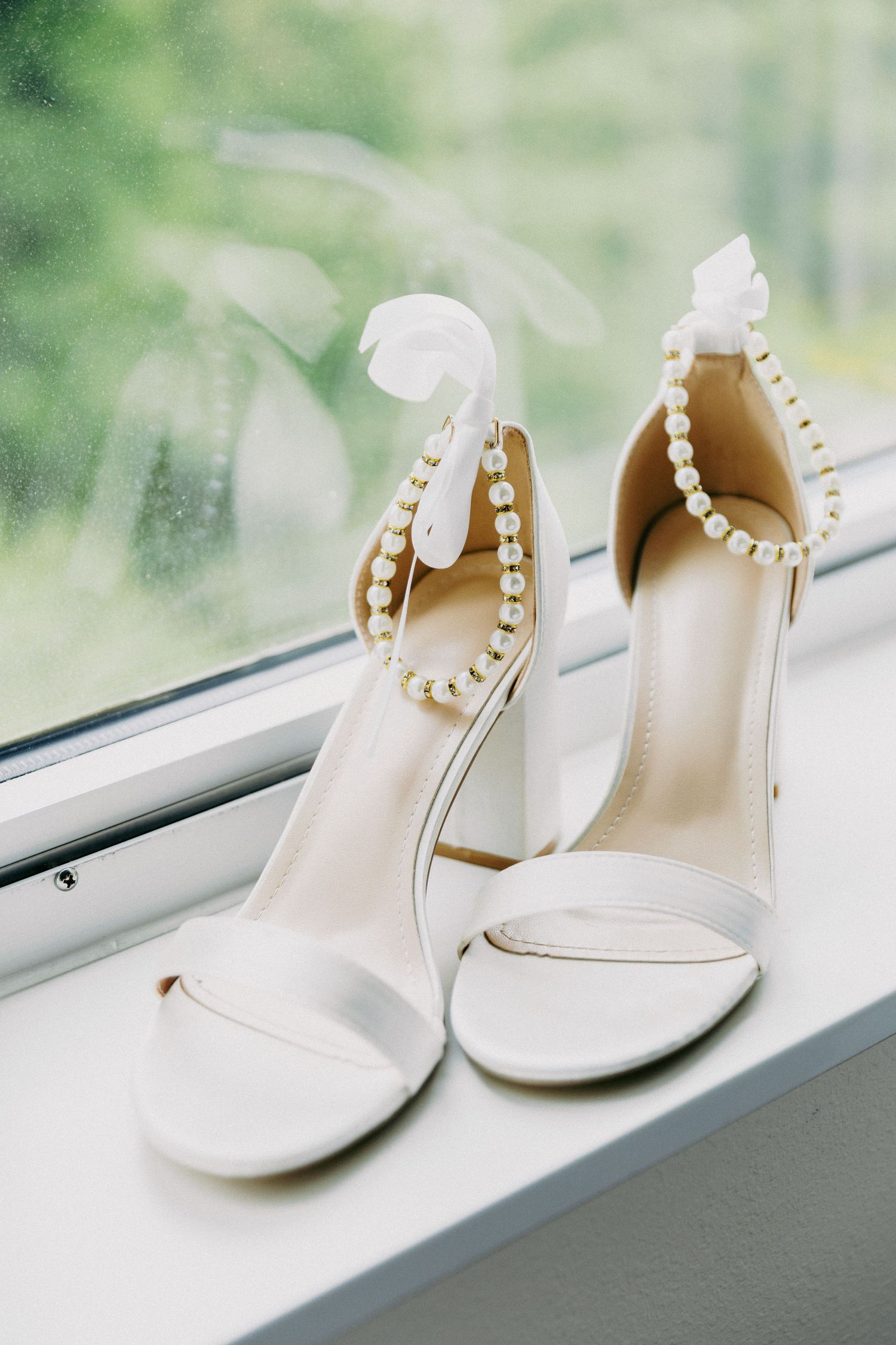 Elegant white bridal heels with pearl ankle straps displayed on a window sill with green garden backdrop.