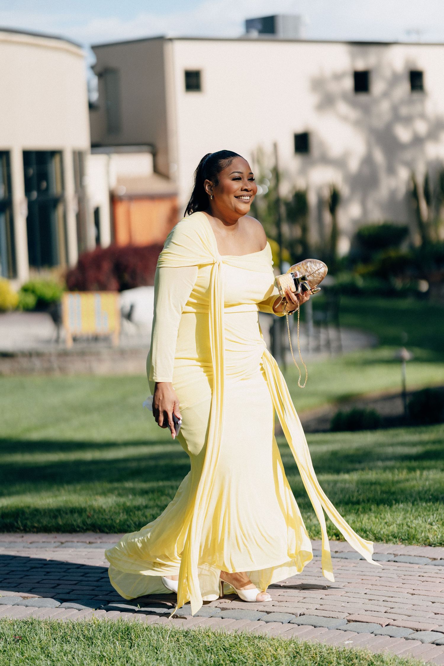 Woman in elegant yellow off-shoulder gown holding clutch purse walks outdoors at wedding venue.