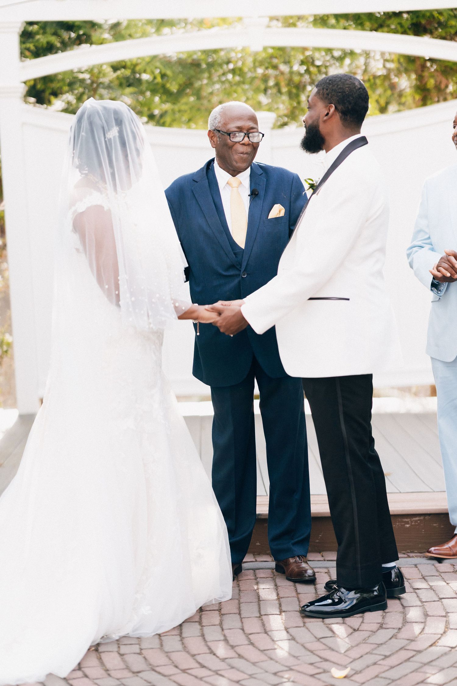 A bride and groom hold hands during an outdoor wedding ceremony while an officiant in a navy suit presides.