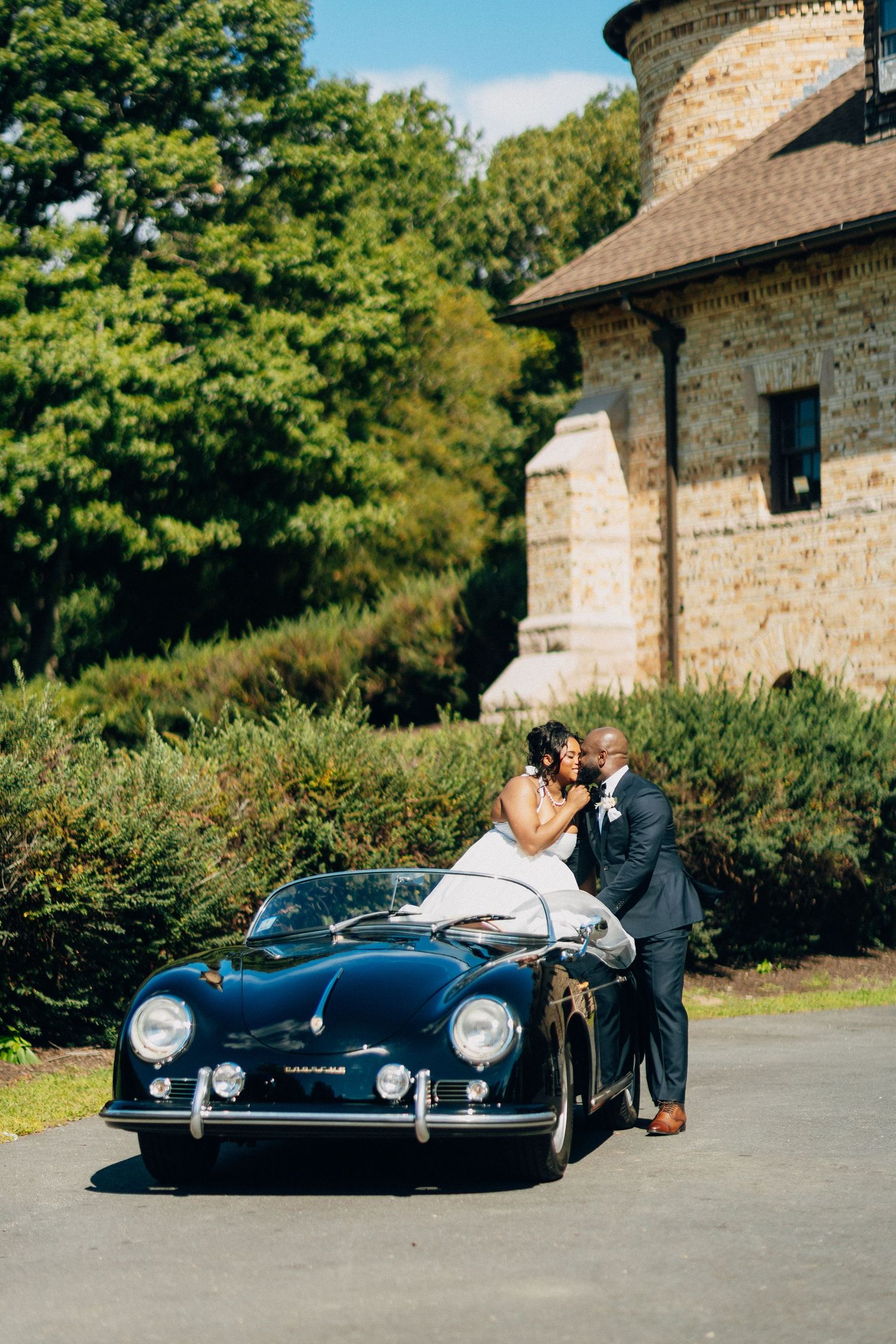 Newlyweds share romantic kiss beside vintage navy blue Porsche 356 speedster at stone venue.