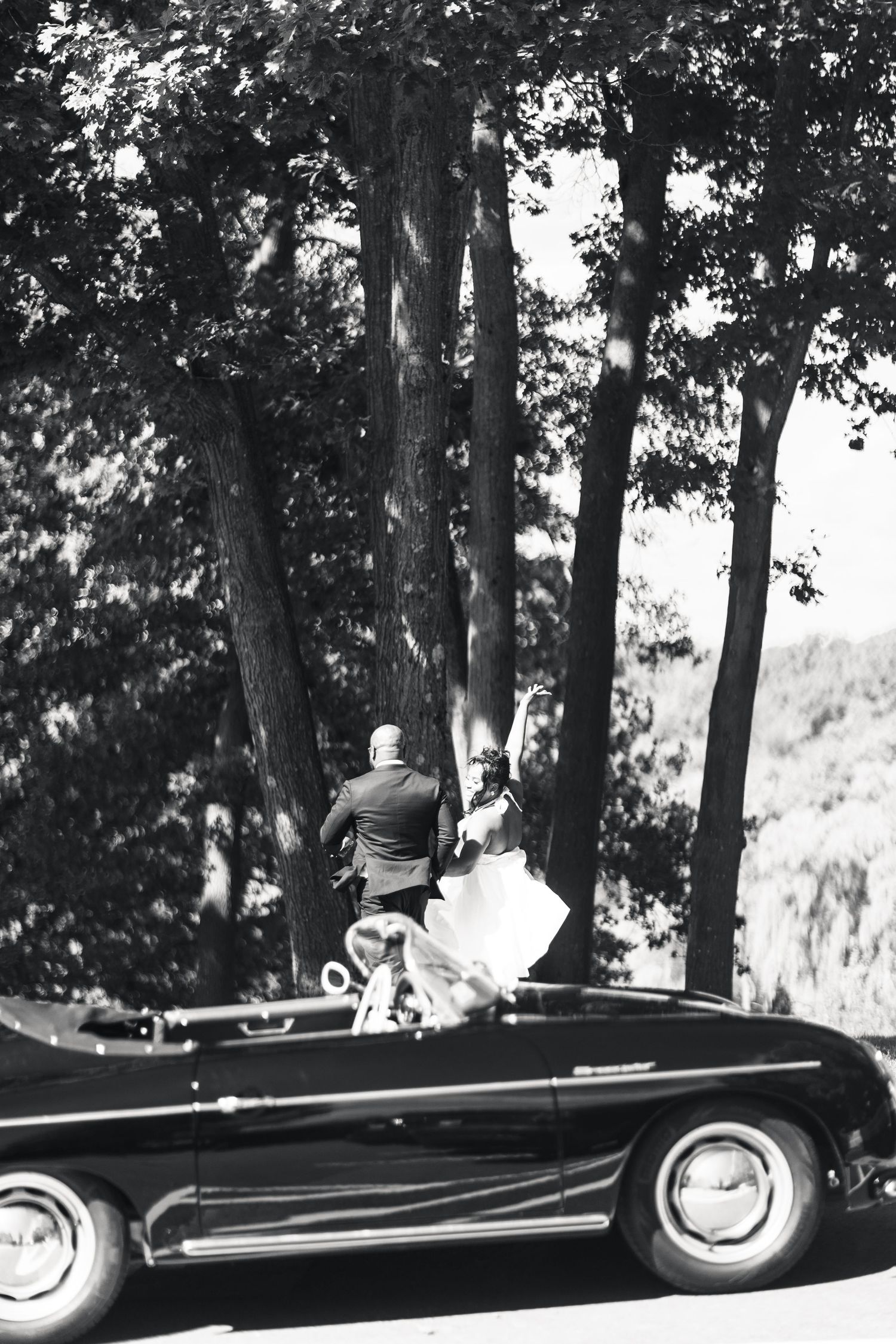 Black and white photo of bride and groom walking past a vintage convertible car under tall trees.