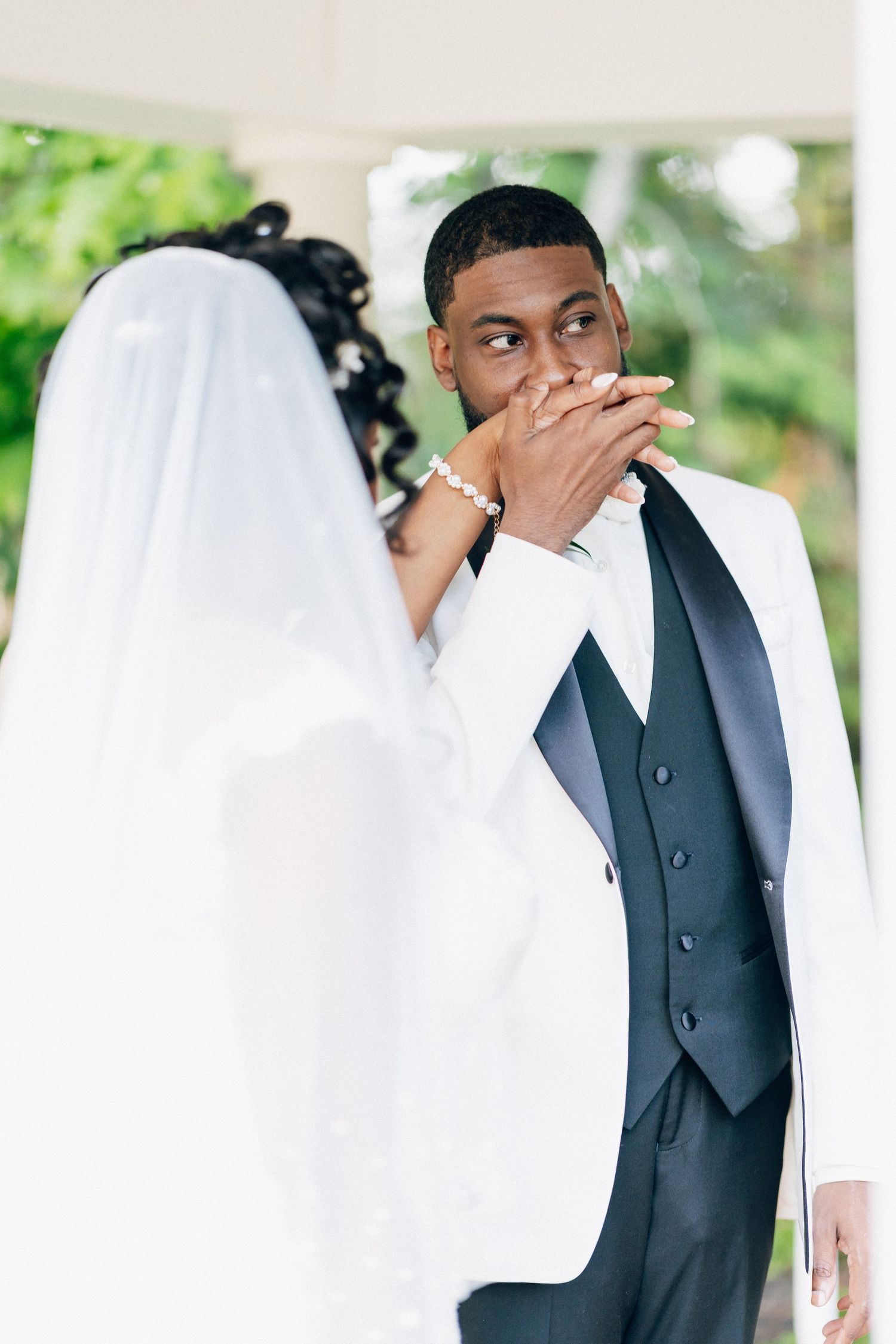 Groom in white tuxedo kisses bride's hand during outdoor wedding ceremony with lush green background.
