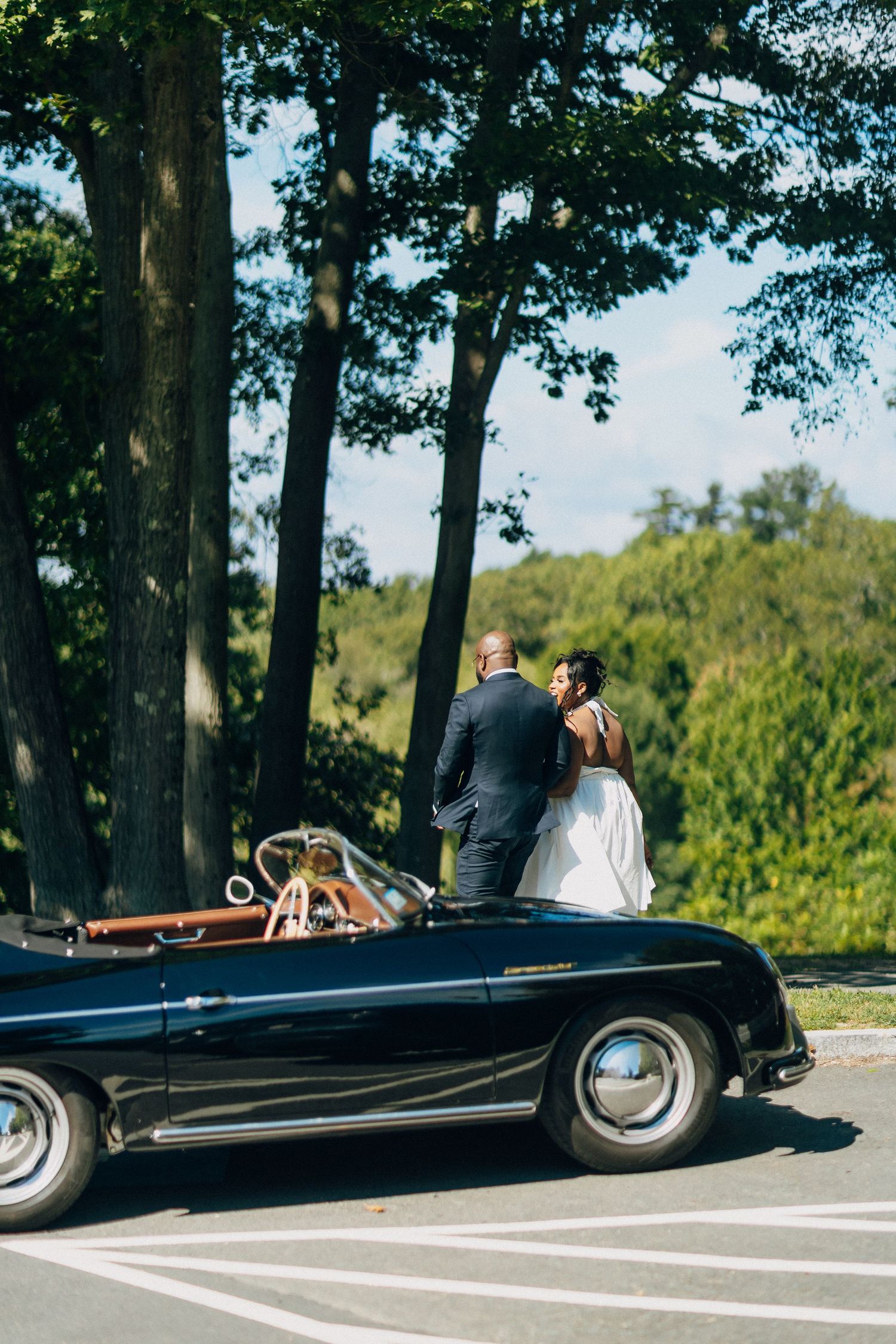Bride and groom share a romantic moment beside a vintage black convertible car under tall trees.