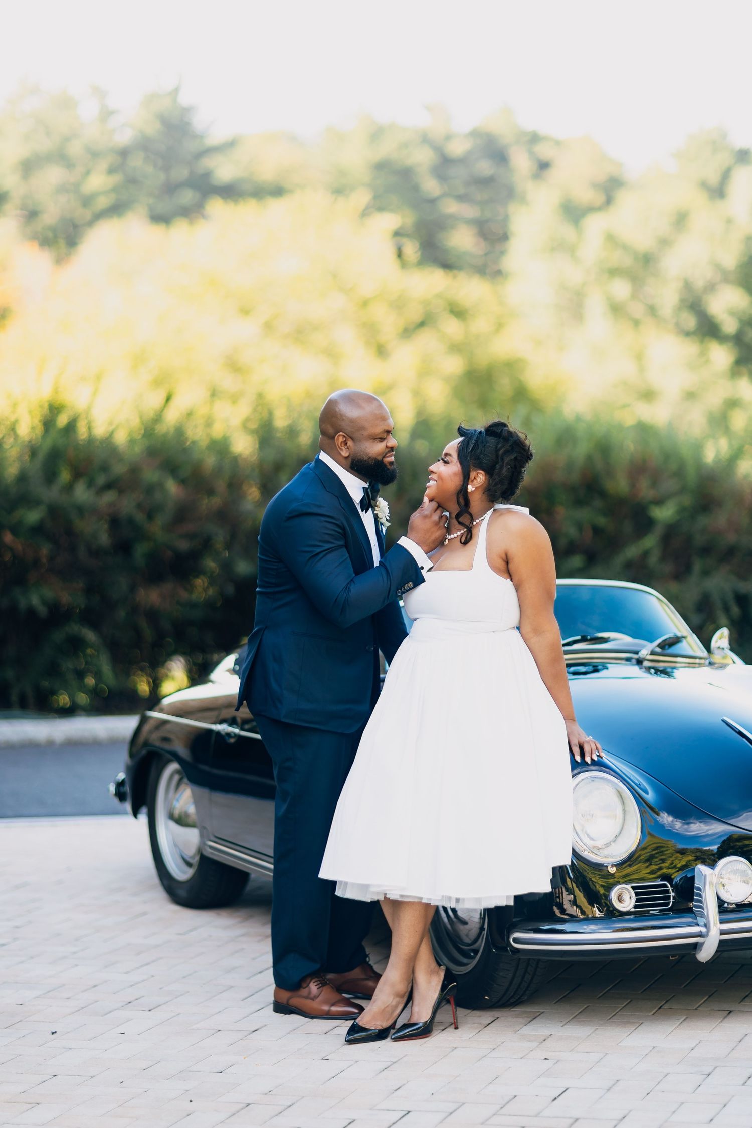 Couple in wedding attire share a romantic moment leaning against a classic black vintage car outdoors.