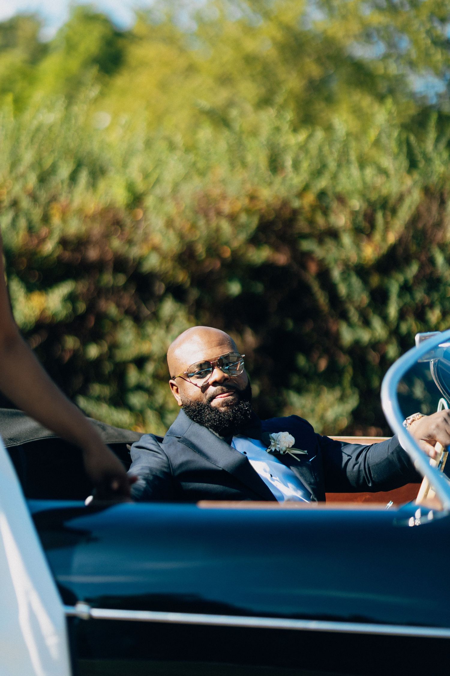 Dapper groom in navy tuxedo sits in vintage convertible wearing stylish sunglasses on wedding day.