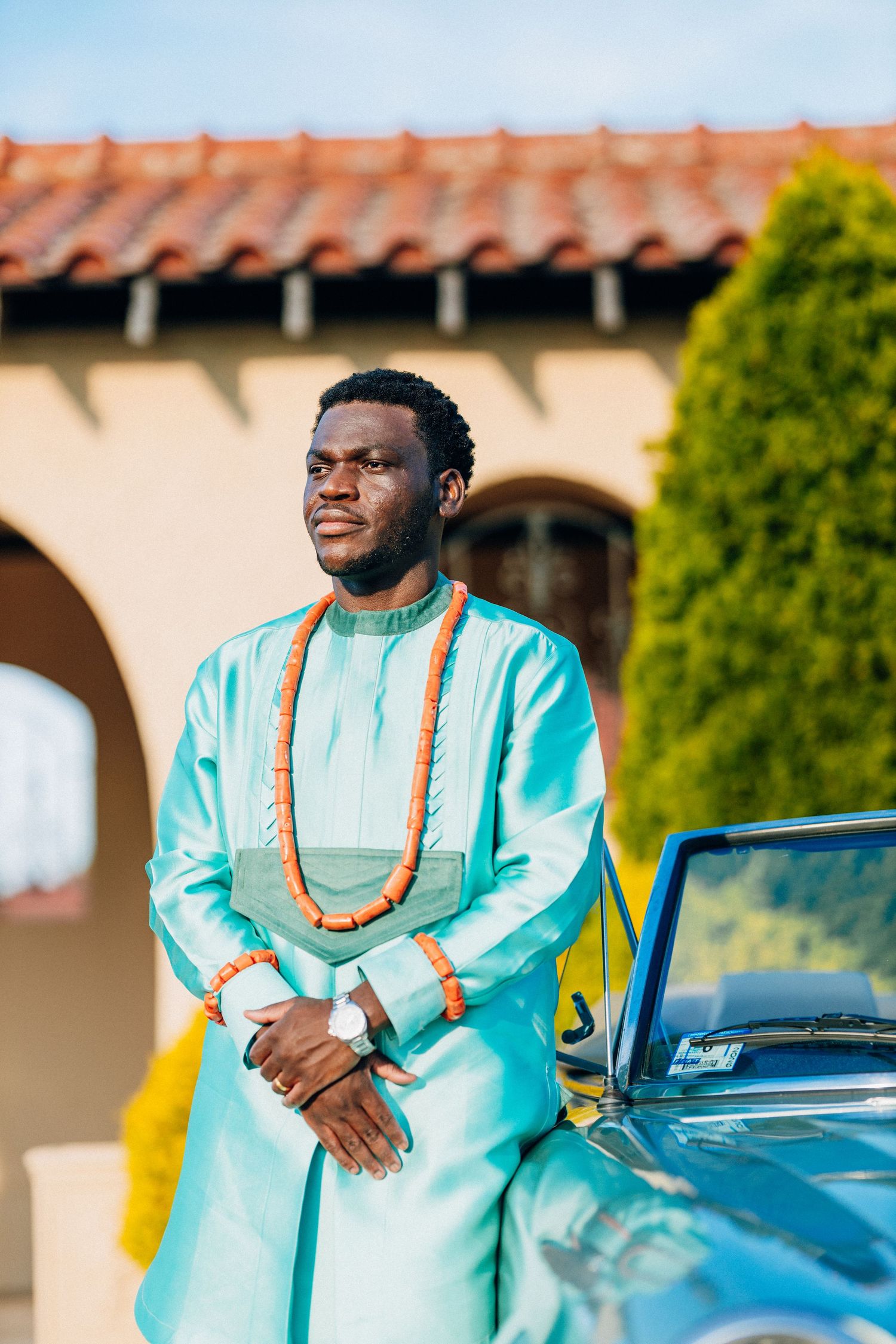 Groom in light blue African traditional attire with coral beads leaning on vintage convertible.