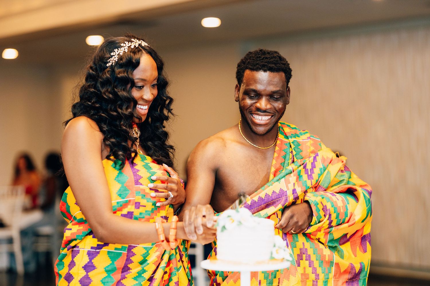 Couple in colorful kente cloth attire cutting wedding cake together at reception.