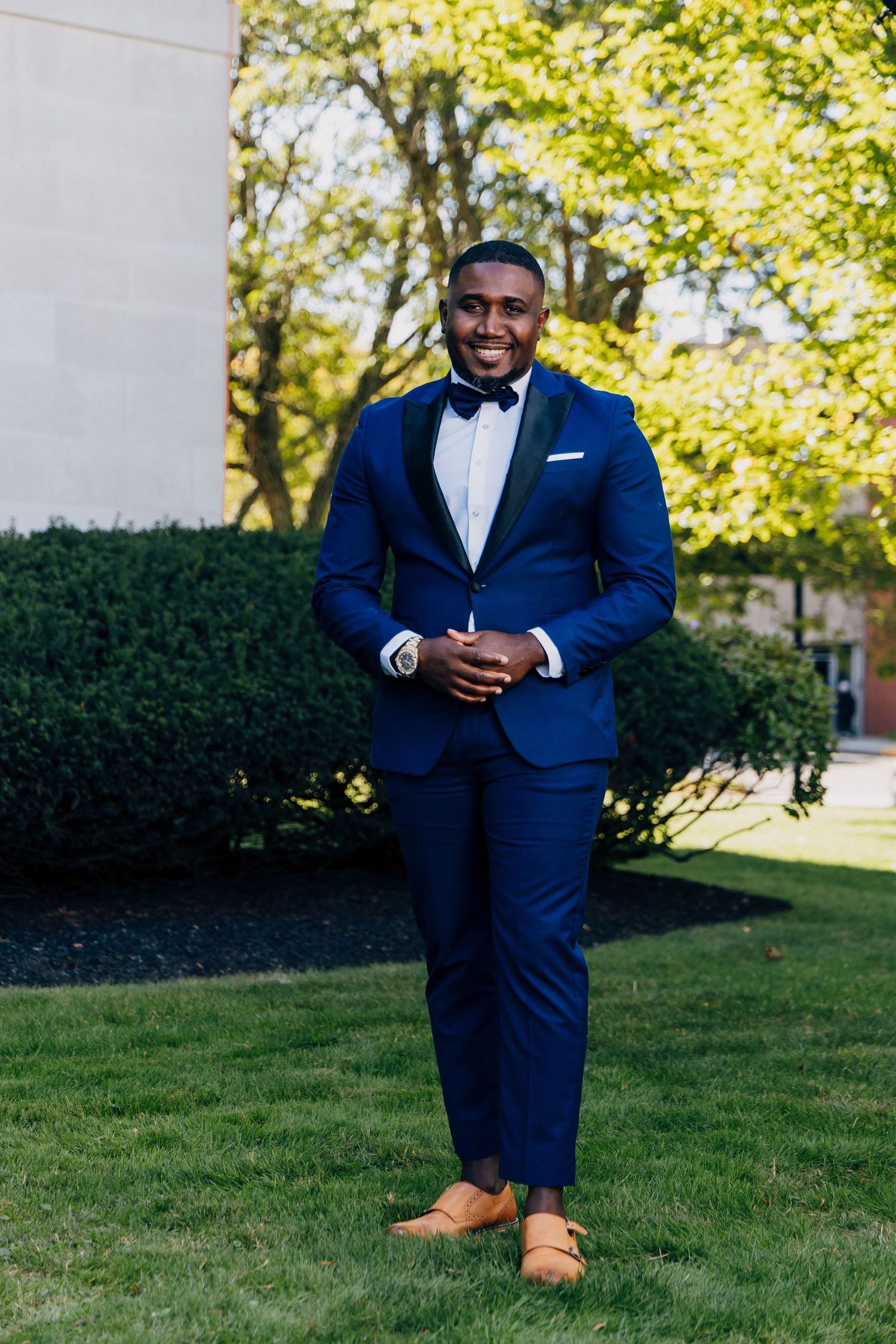 Handsome groom in navy blue tuxedo with bow tie and tan shoes standing on green lawn outdoors.