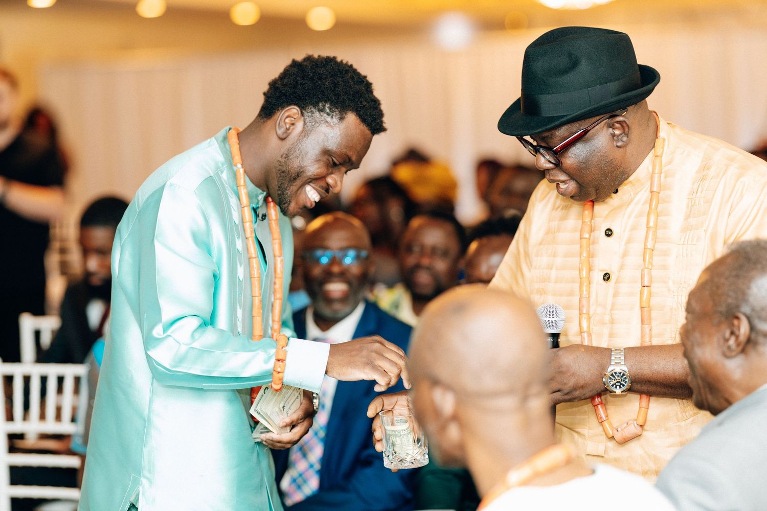 Two men in traditional African attire share a joyful moment at a formal event with guests seated in background.