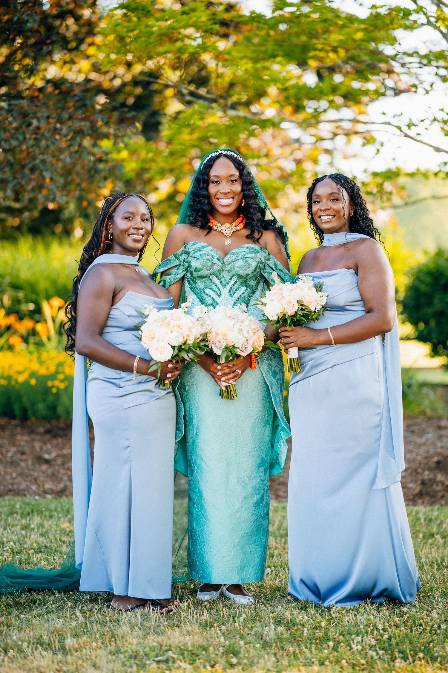 Bride in teal gown with two bridesmaids in blue dresses holding white bouquets outdoors.