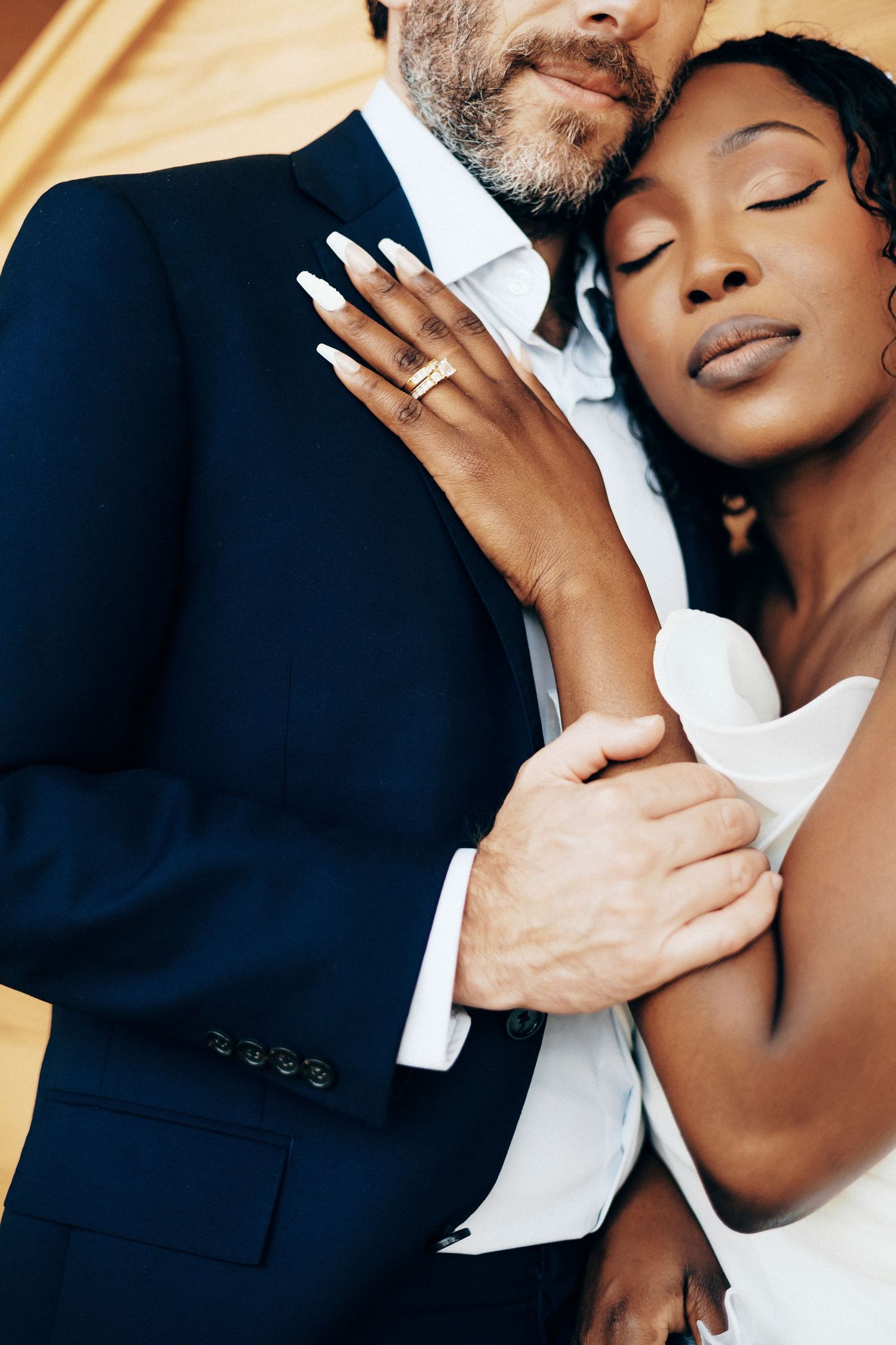 Close-up of interracial wedding couple embracing, bride in white dress showing gold ring, groom in navy suit.