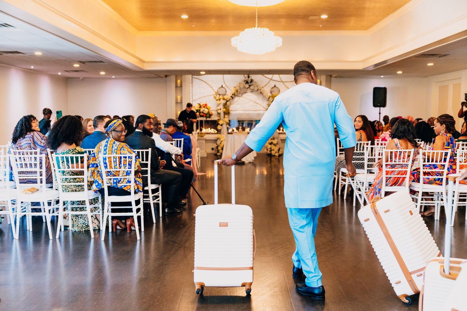 Man in light blue suit pulling white luggage at an elegant wedding reception with white chairs and floral arch.