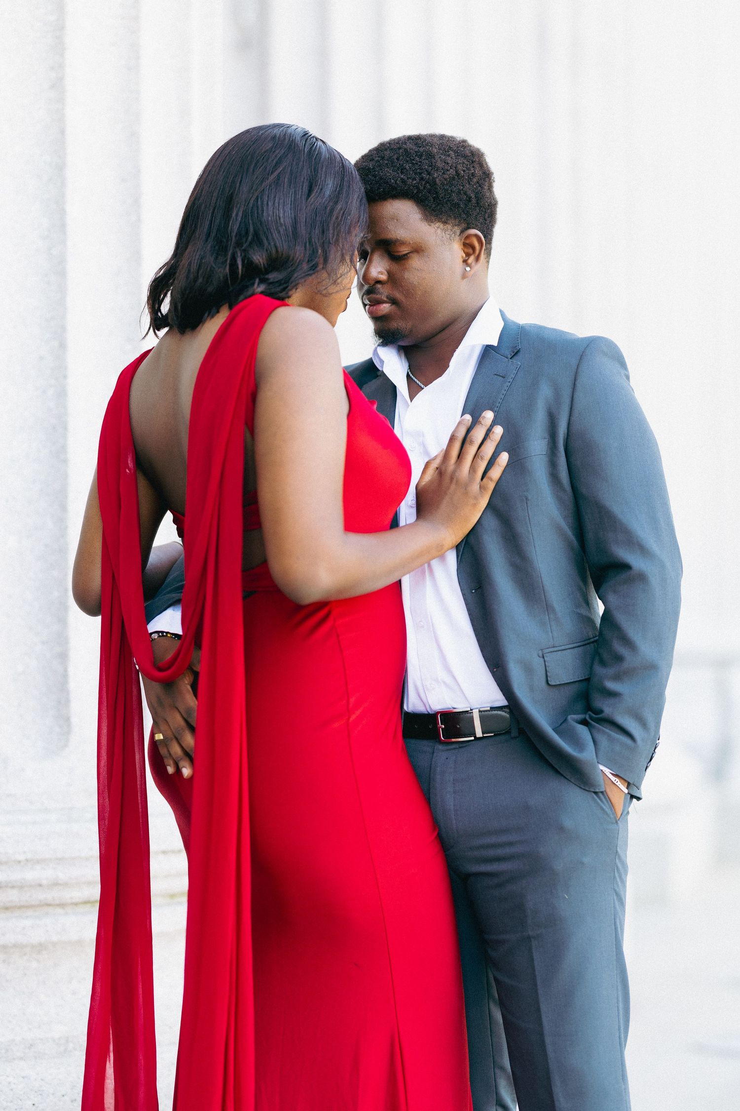 A couple in elegant attire, woman in red gown and man in gray suit, share an intimate moment by white columns.