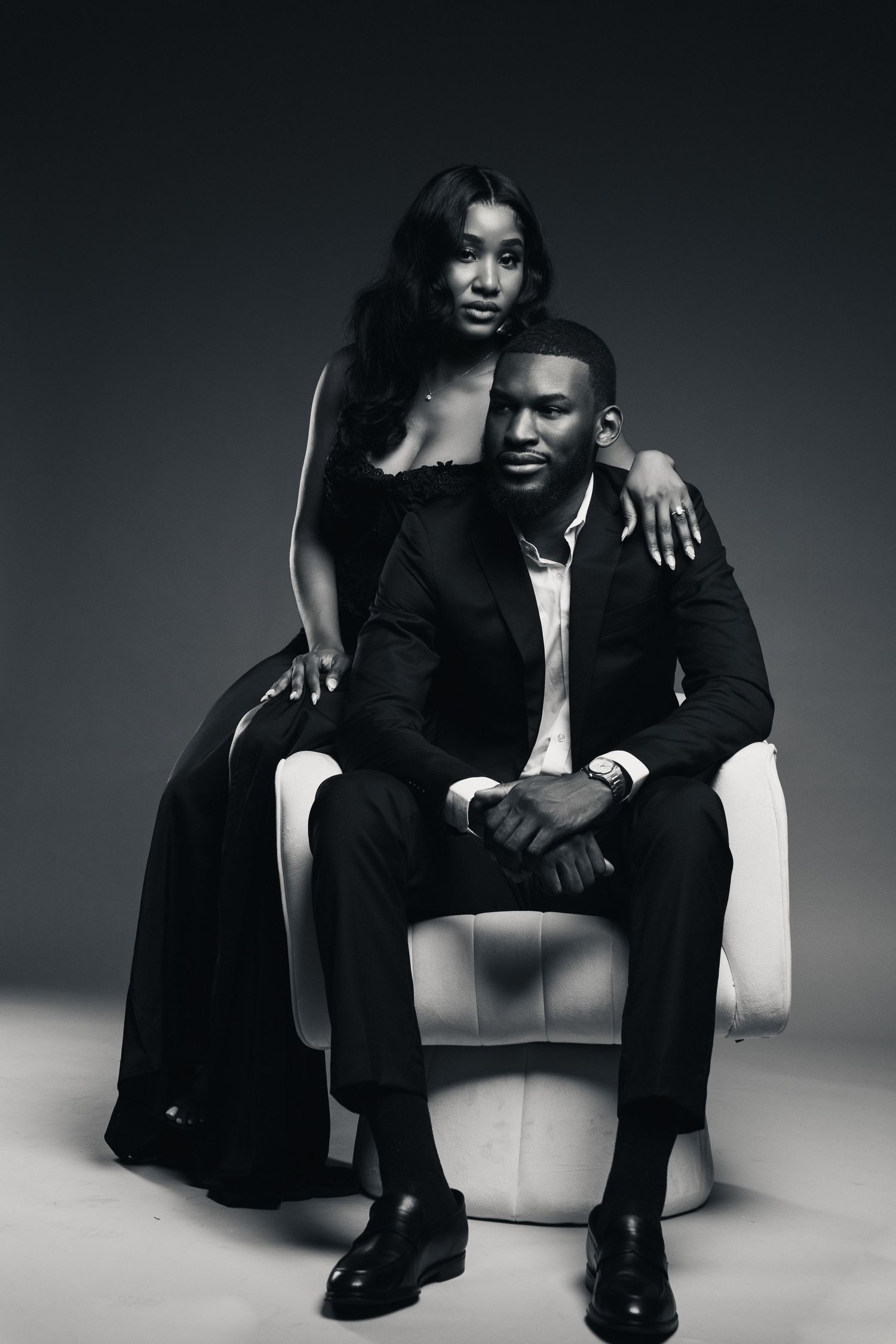 Black and white portrait of an elegant couple, man in suit seated in chair, woman in evening gown standing beside him.