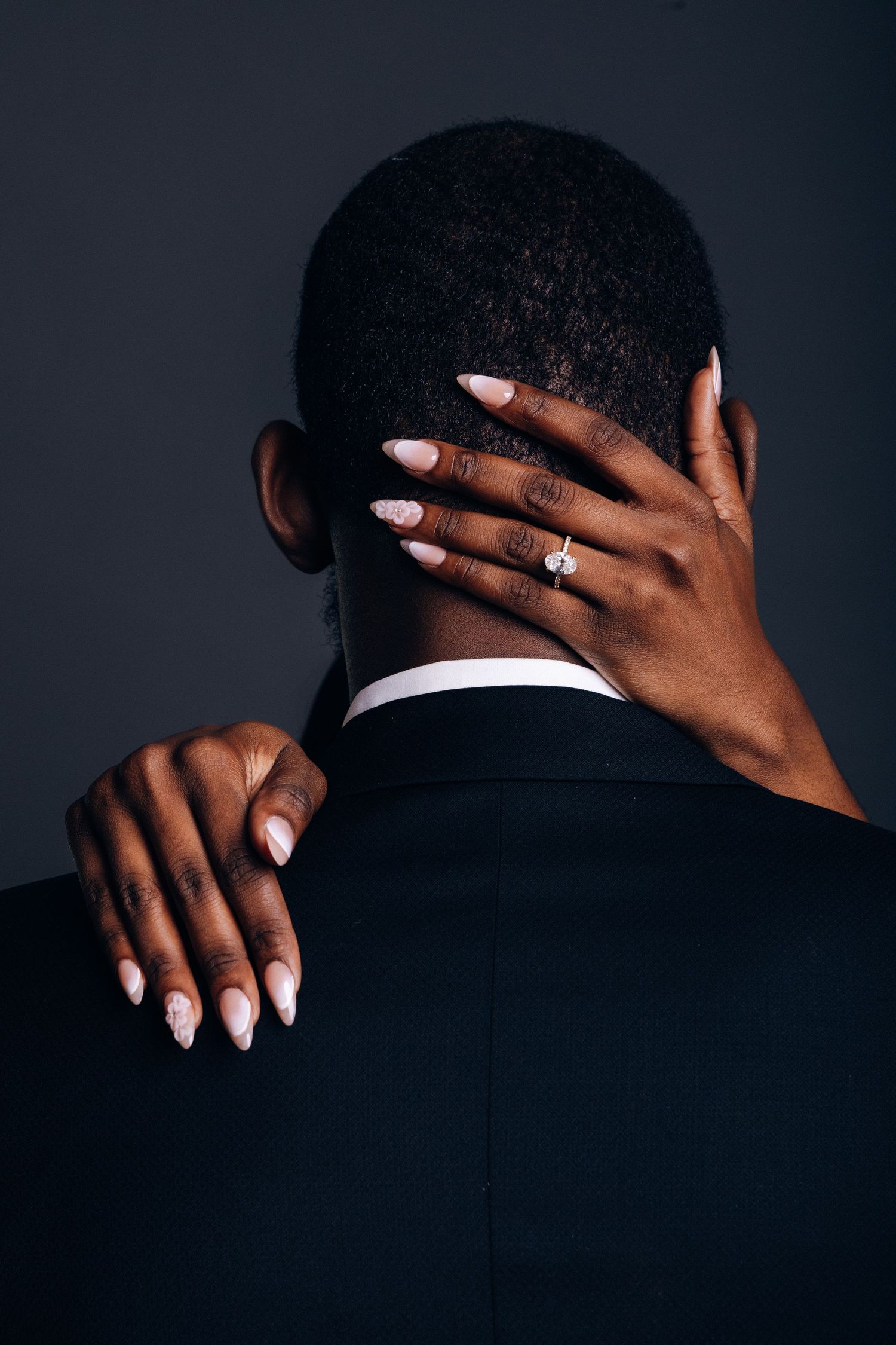A woman with pink nails and a diamond engagement ring embraces a man in a suit from behind against a dark background.