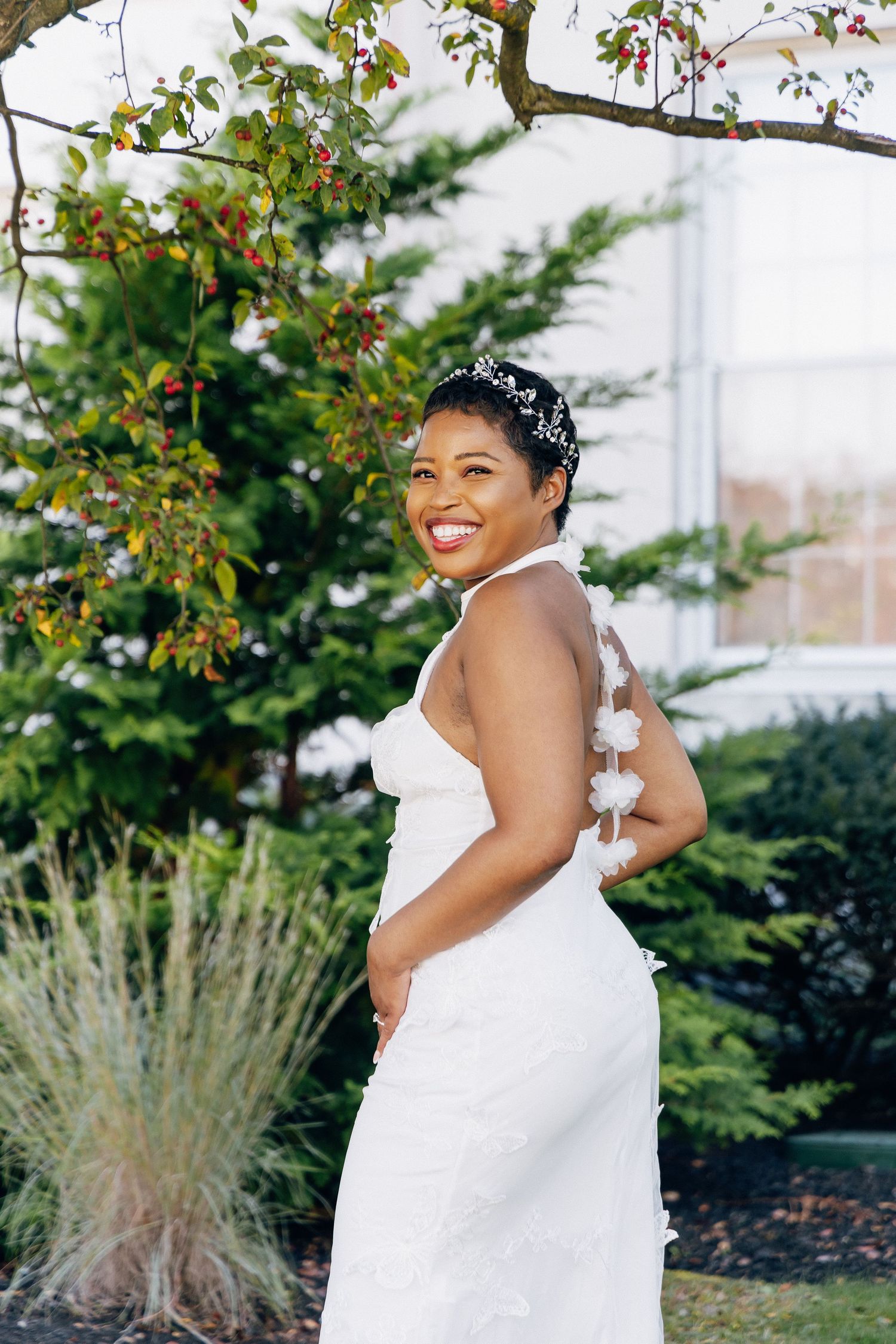 Bride in backless white halter gown with floral details smiling outdoors near blooming tree.