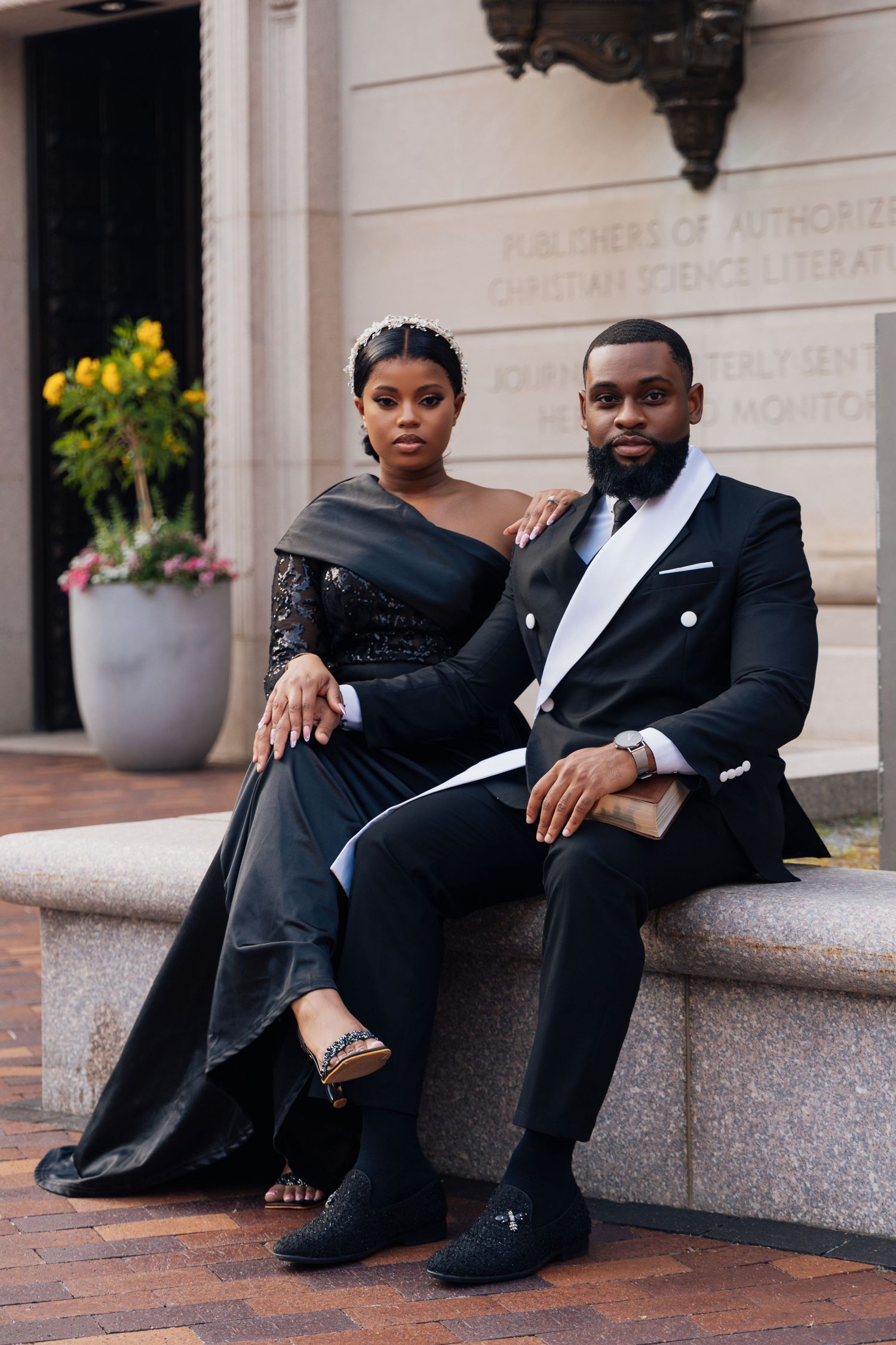 Elegant couple in matching black formal attire sitting on stone ledge outside a classic building.