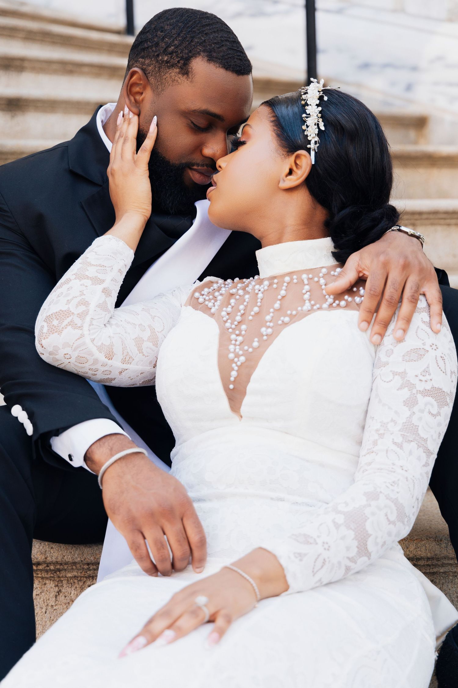 A Black bride and groom share a romantic moment on stone steps, bride in lace gown, groom in black suit.