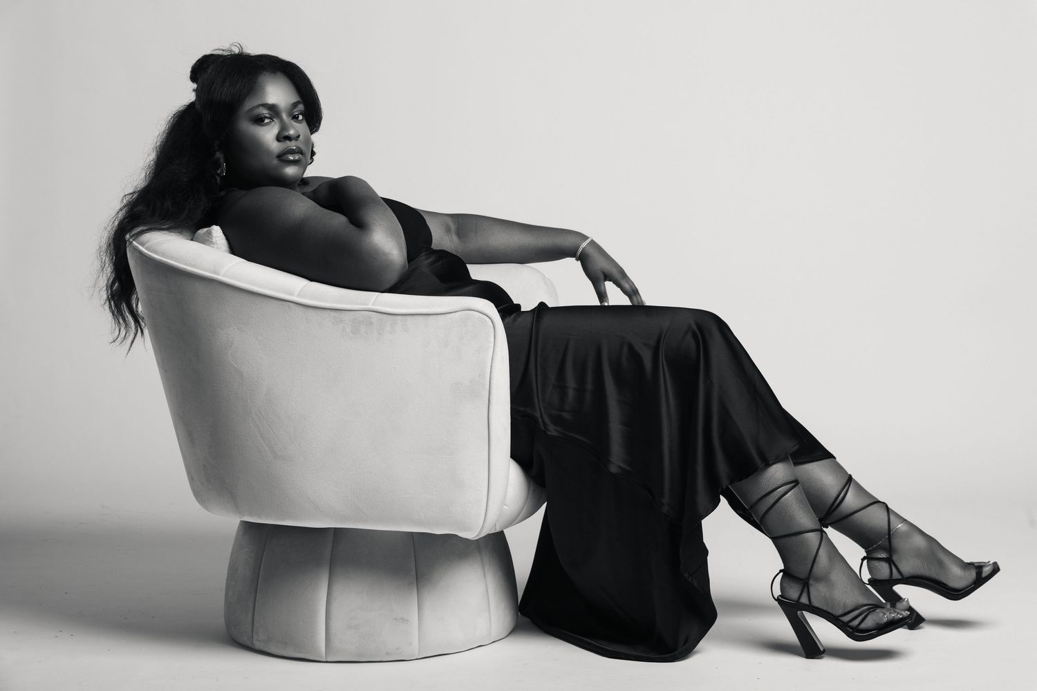 Black and white photo of a woman in elegant black outfit reclining gracefully in a white velvet chair with strappy heels.