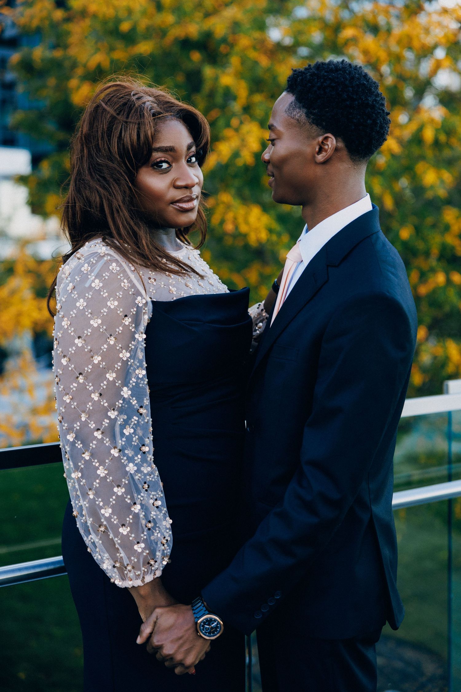 Elegant couple in formal attire holding hands outdoors, woman in lace dress, man in navy suit, autumn trees background.