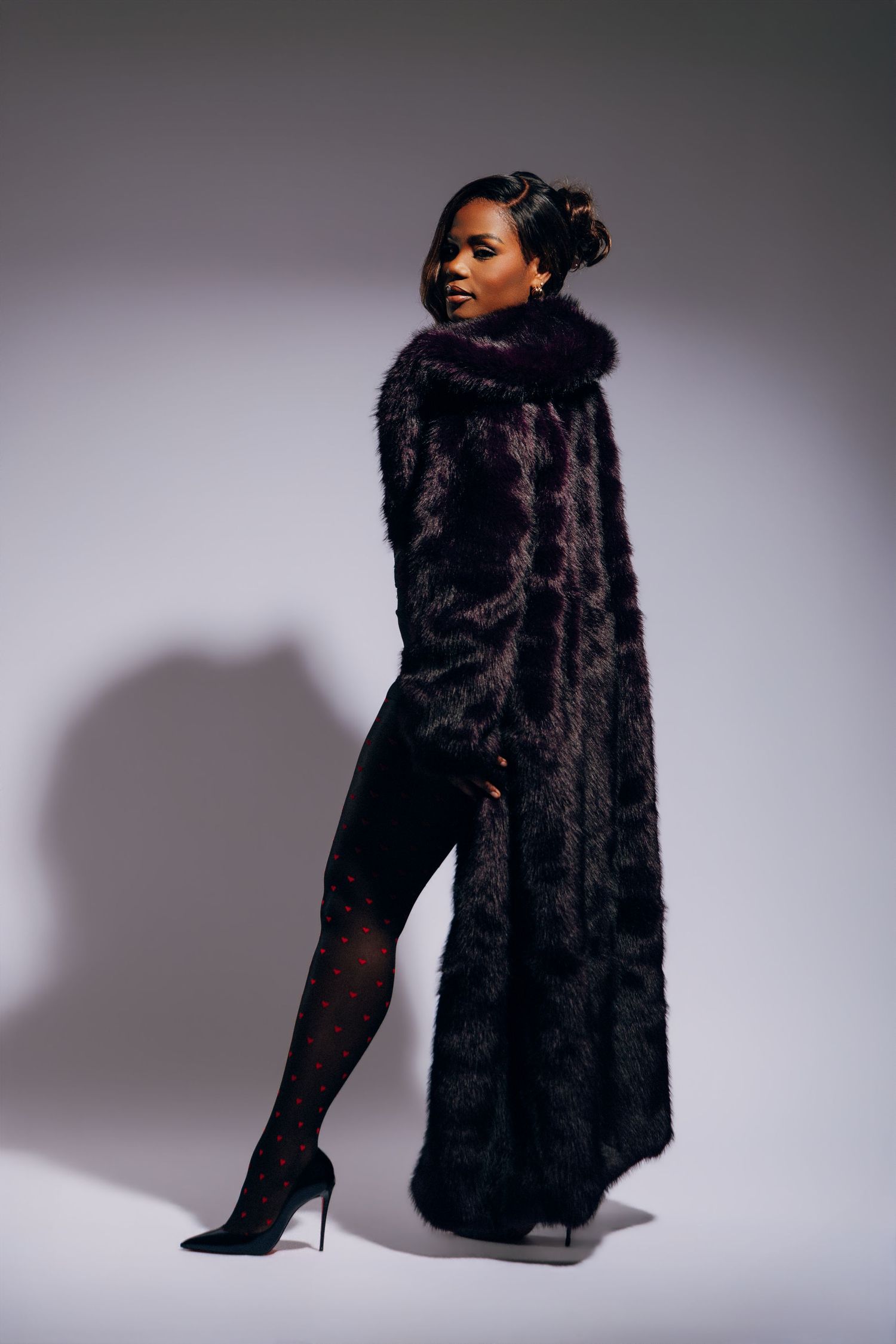Woman in long dark fur coat and heels posing elegantly against a light gray studio background.