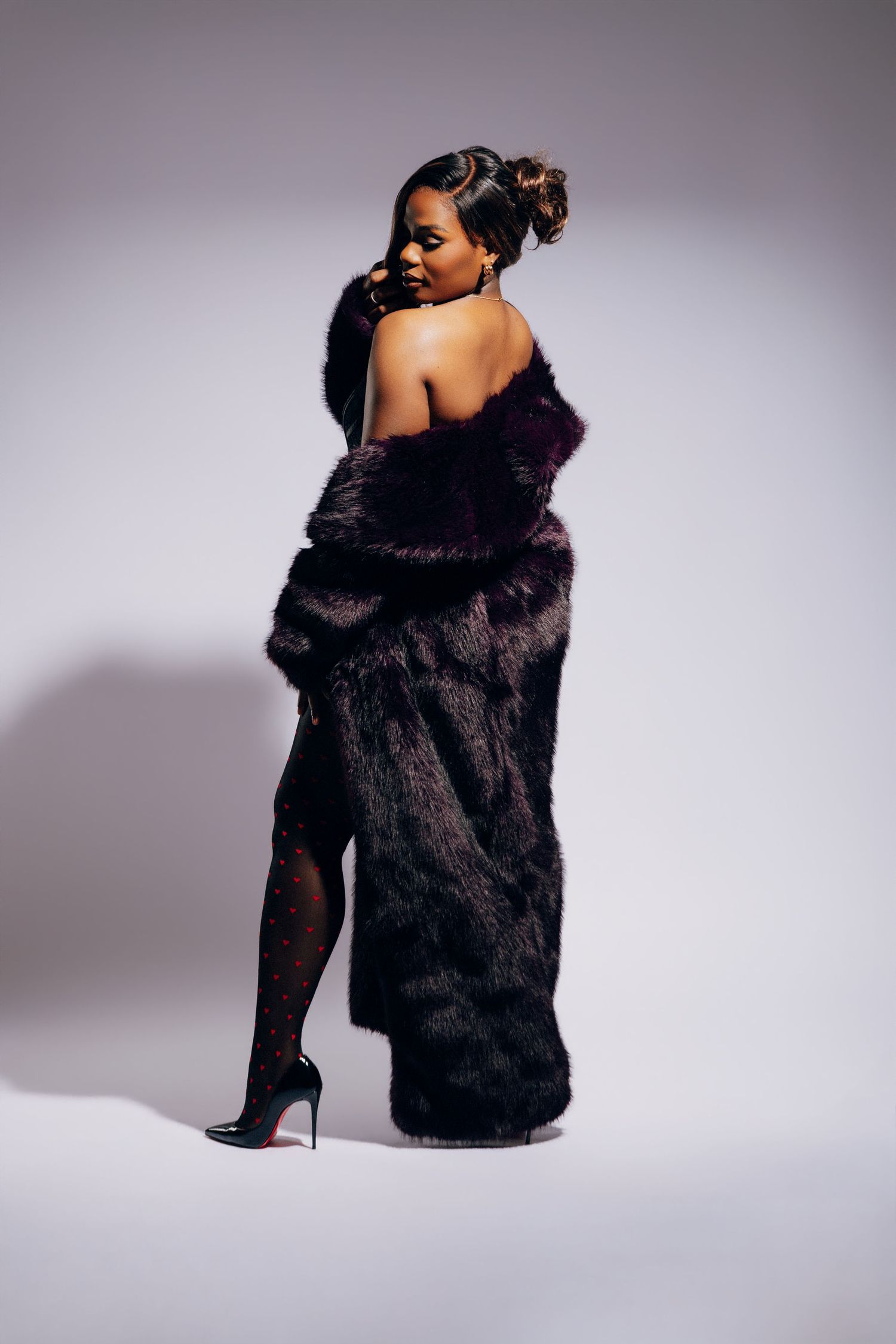 A woman in a dark fur coat and heels poses elegantly in a studio, showing off her stylish outfit.