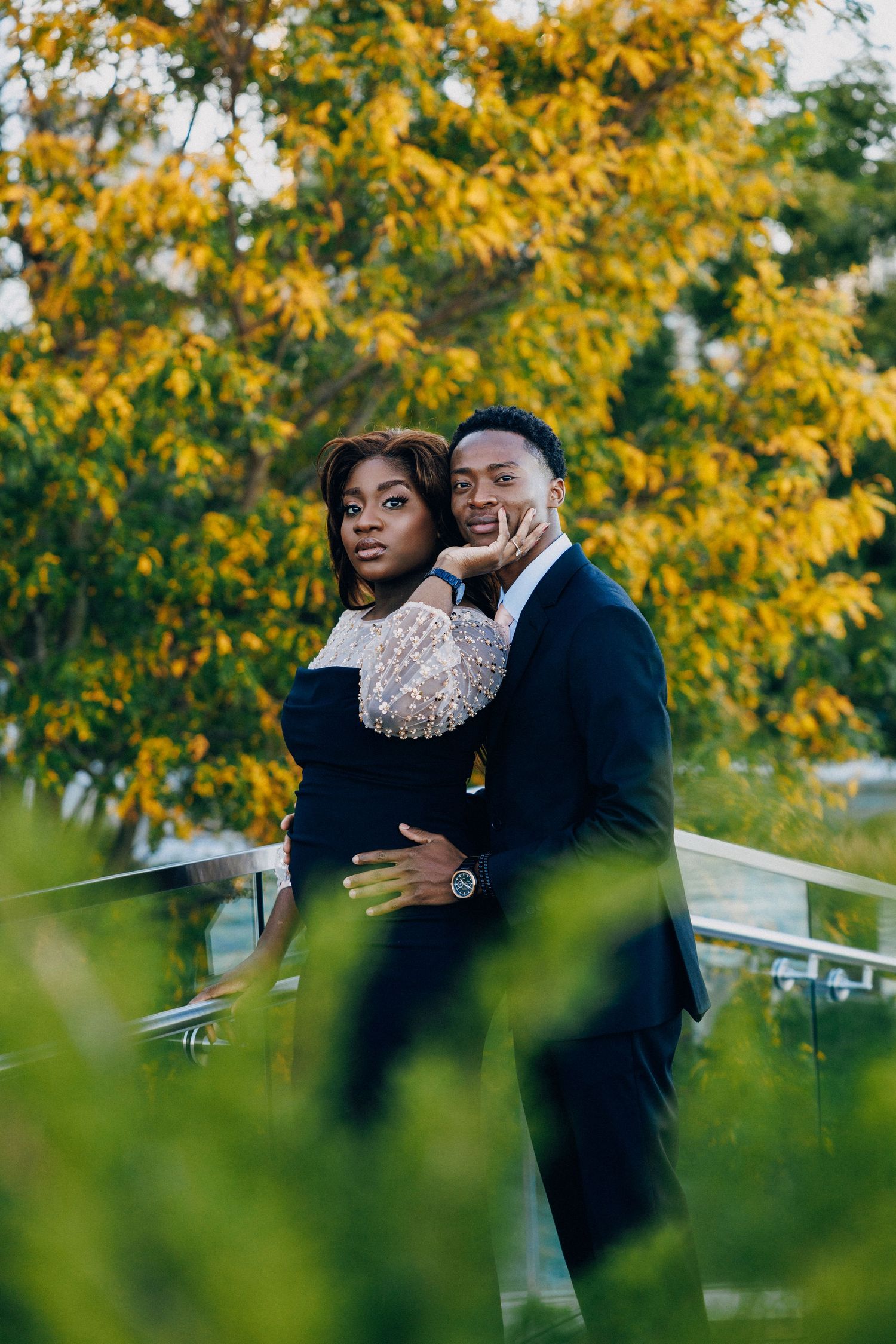 Elegant Black couple embracing outdoors, woman in pearl-embellished dress, man in suit, surrounded by autumn foliage.