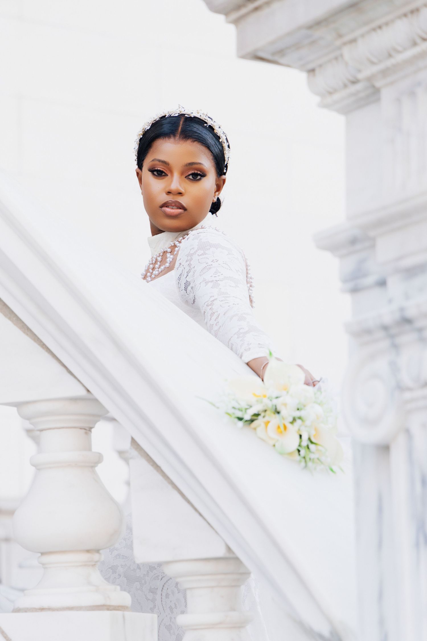 Elegant Black bride in lace gown holding white bouquet on marble staircase with ornate white columns.