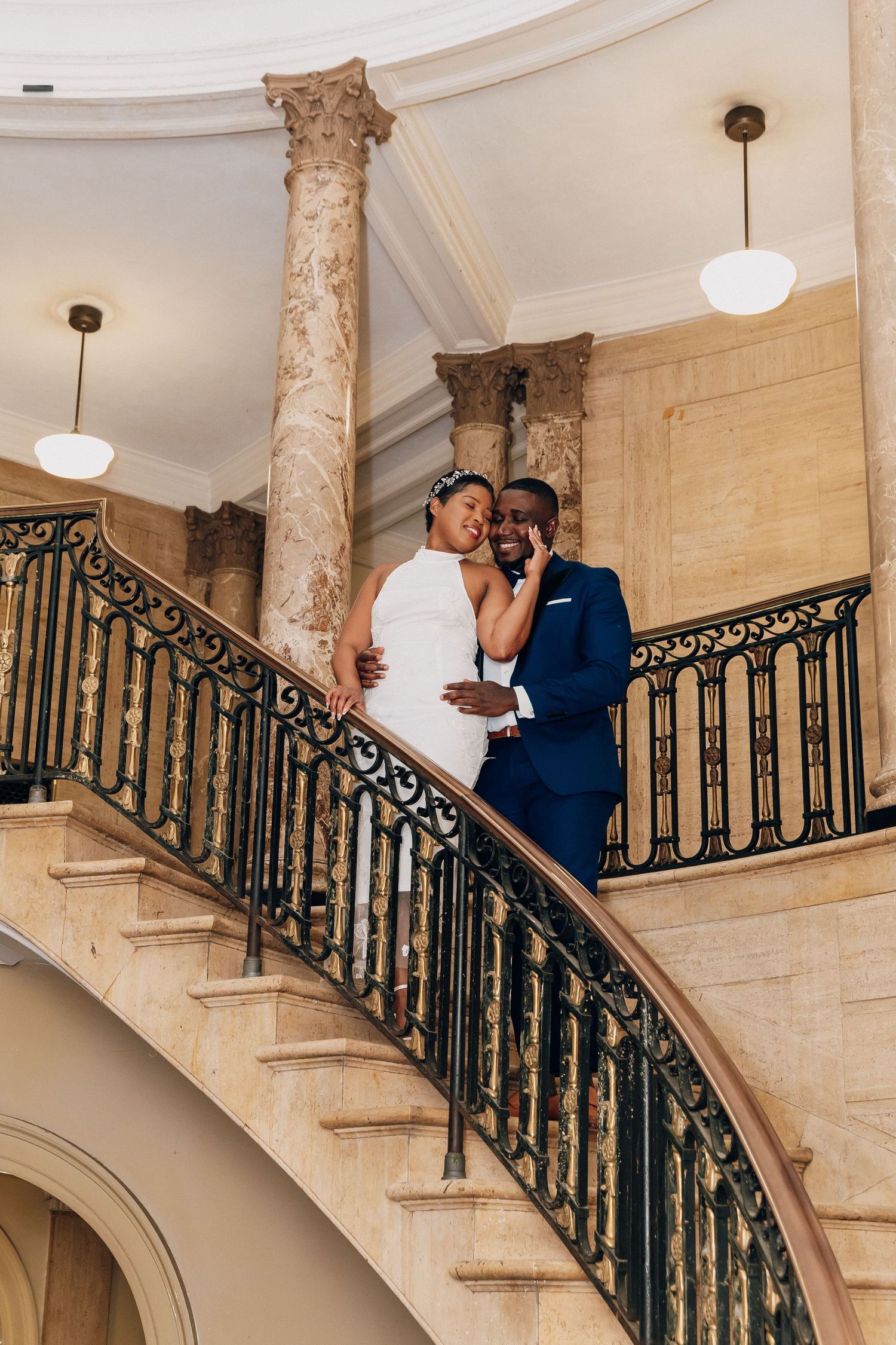 Newlyweds embrace on curved grand staircase with ornate iron railing in elegant marble wedding venue.