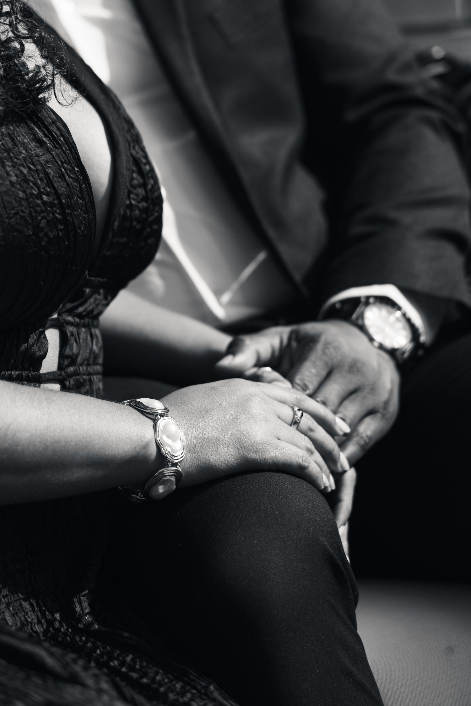 Black and white photo of a couple holding hands, both wearing watches, woman with ring, dressed elegantly.