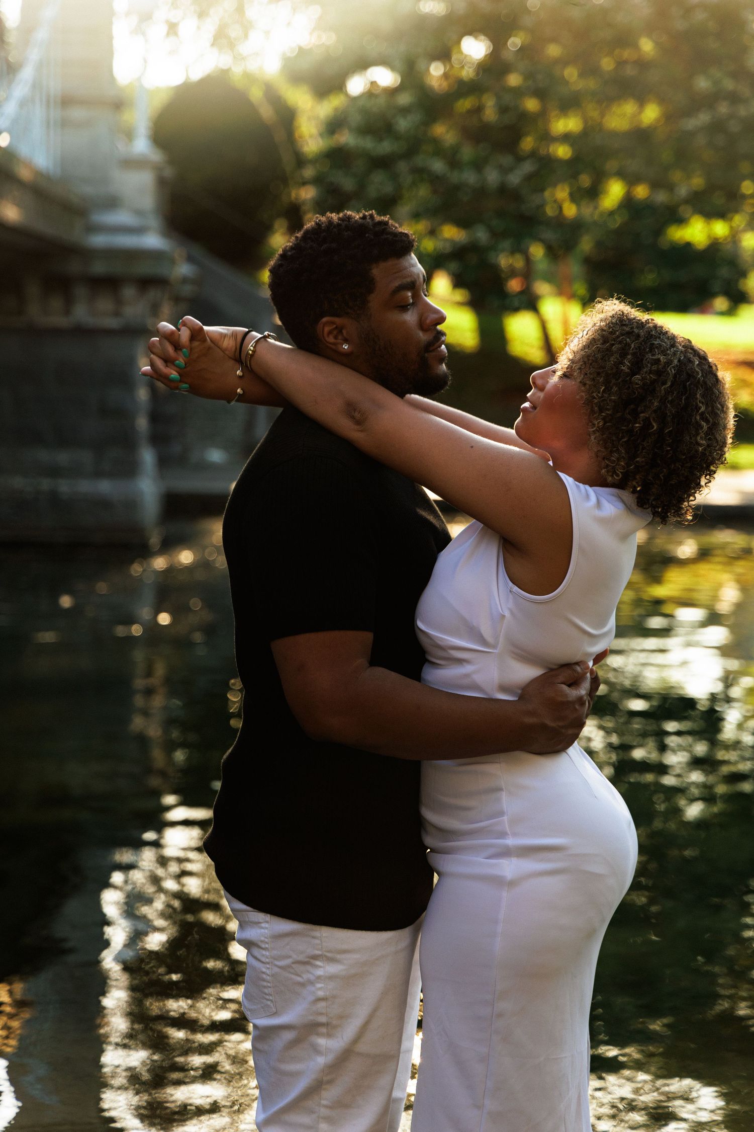 A loving couple hugs near a fountain in a sun-drenched park, the woman in white gazing up at her partner affectionately.