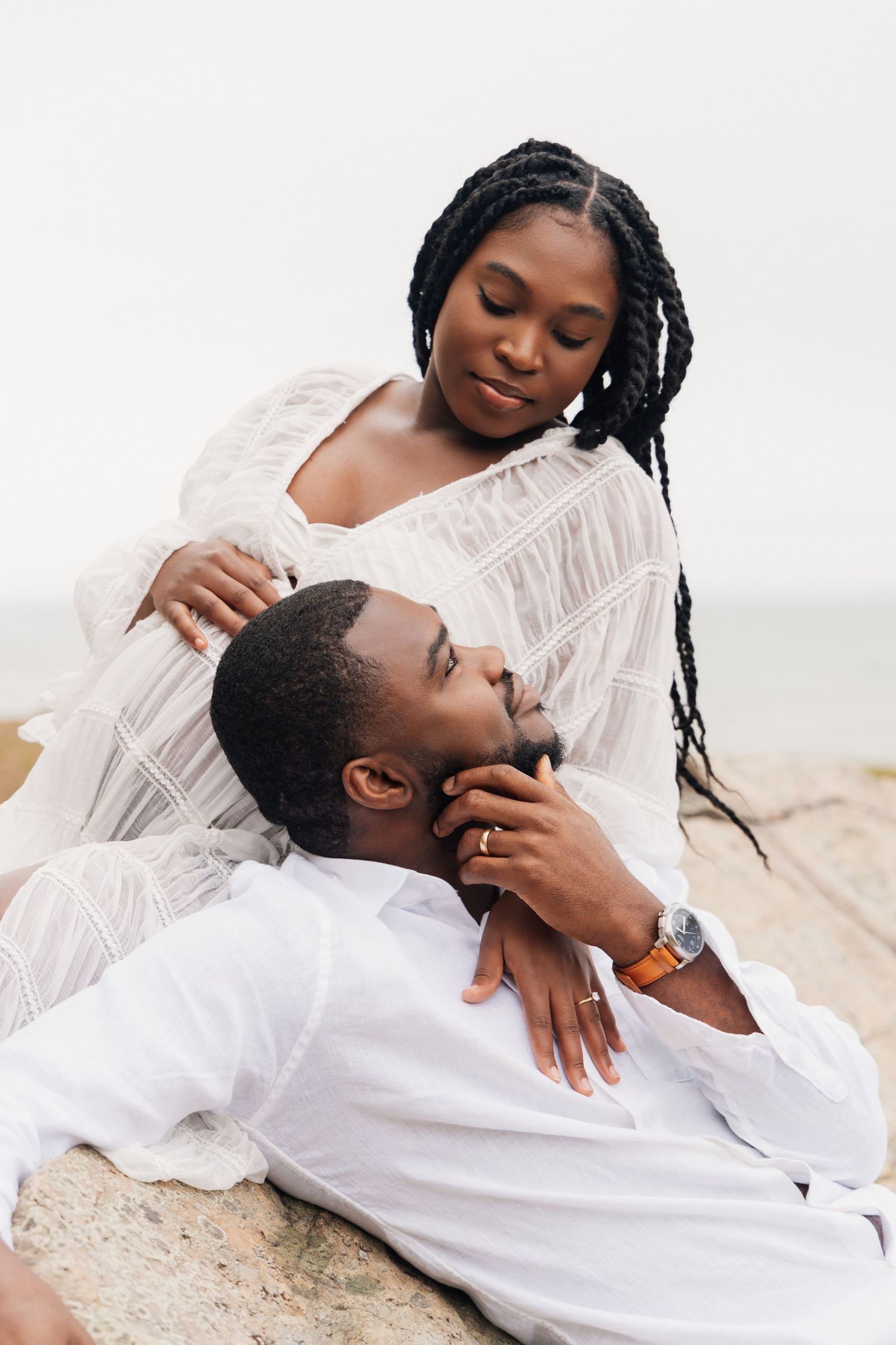 A Black couple shares an intimate moment on rocks by the ocean, the woman cradling the mans face tenderly.