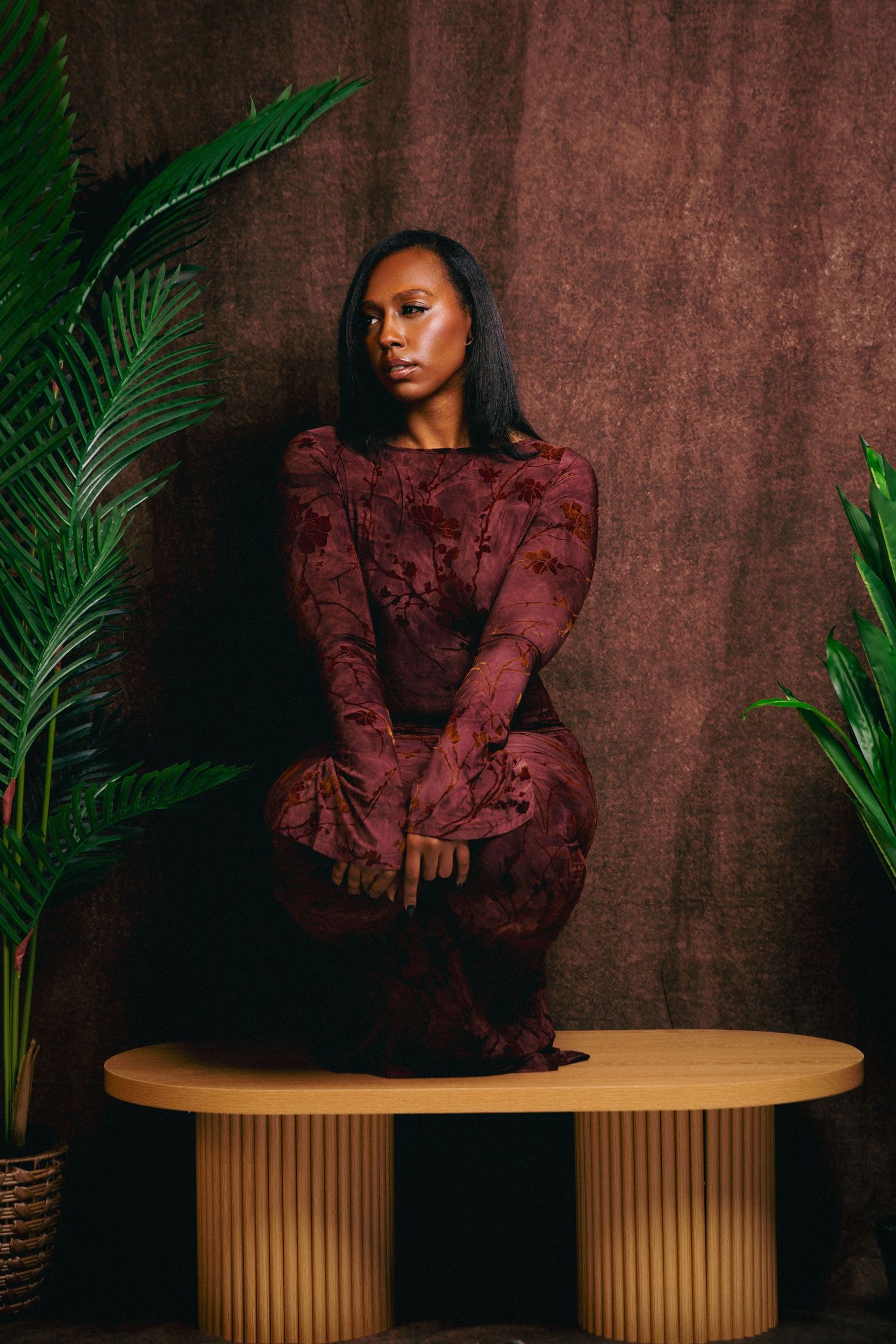 Woman in burgundy floral dress sitting elegantly on oval wooden table.