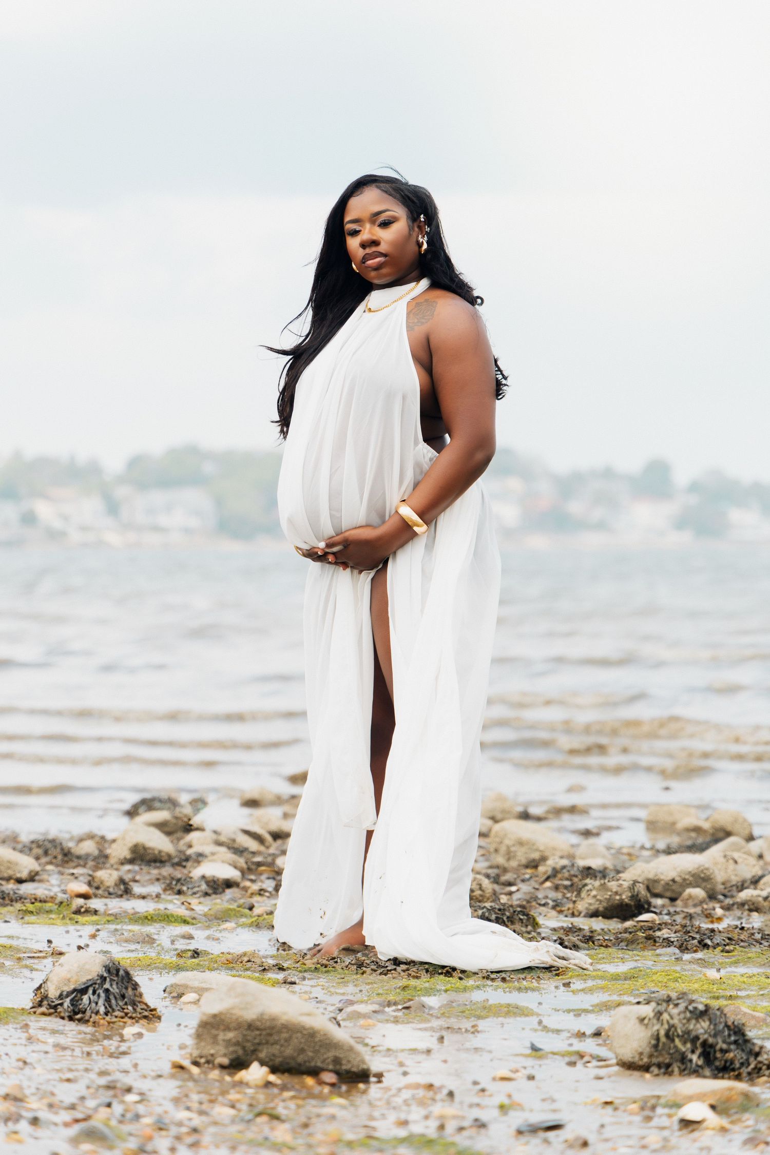 Pregnant woman in white halter dress standing on rocky shoreline with ocean and town in background.
