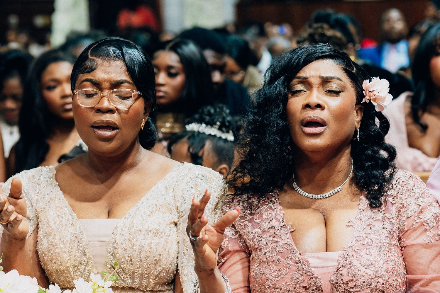 Two women in elegant pink gowns singing together at a wedding ceremony surrounded by guests.