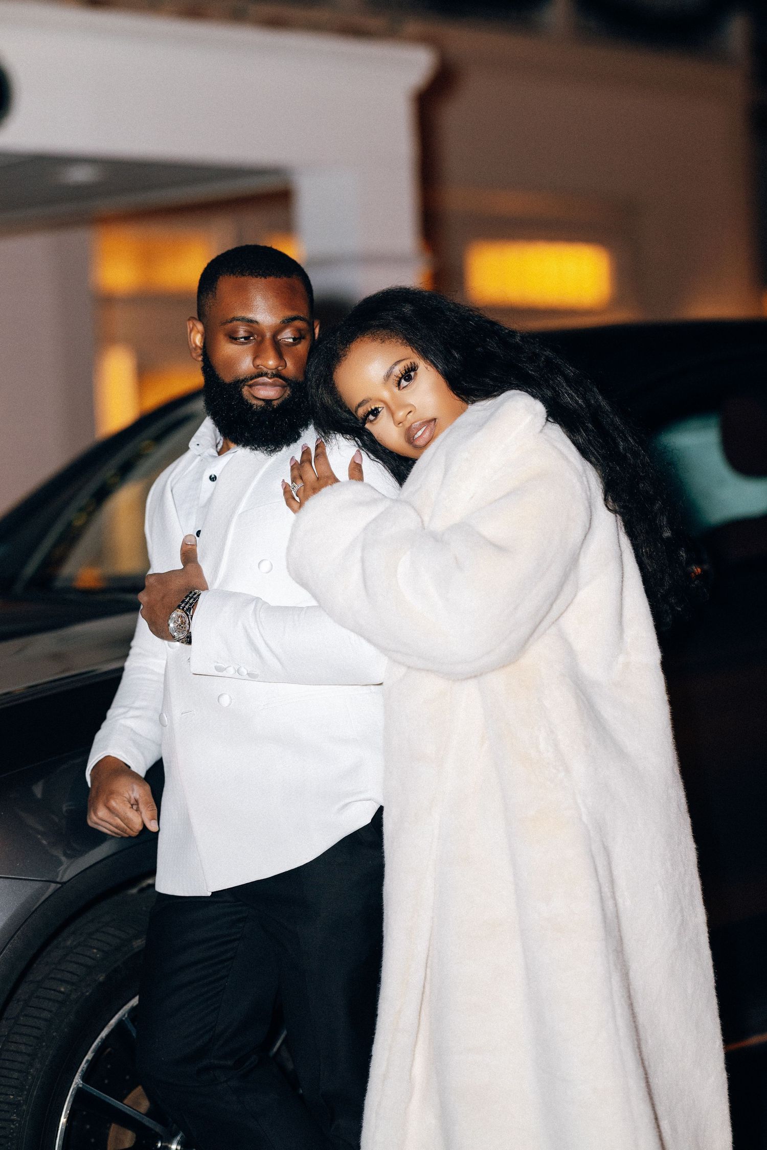 Newlywed couple posing together outdoors at night, bride wearing a white fur coat beside a luxury vehicle.