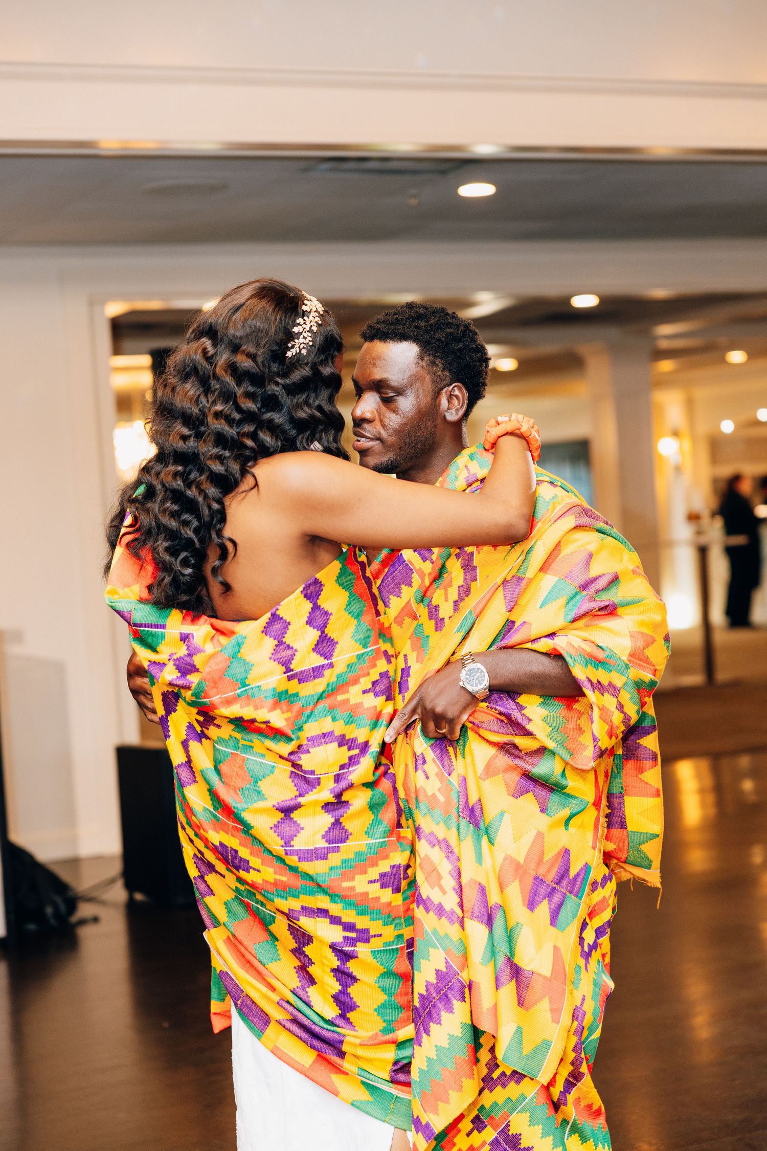 Couple dancing in vibrant African kente cloth attire at an elegant indoor wedding reception.