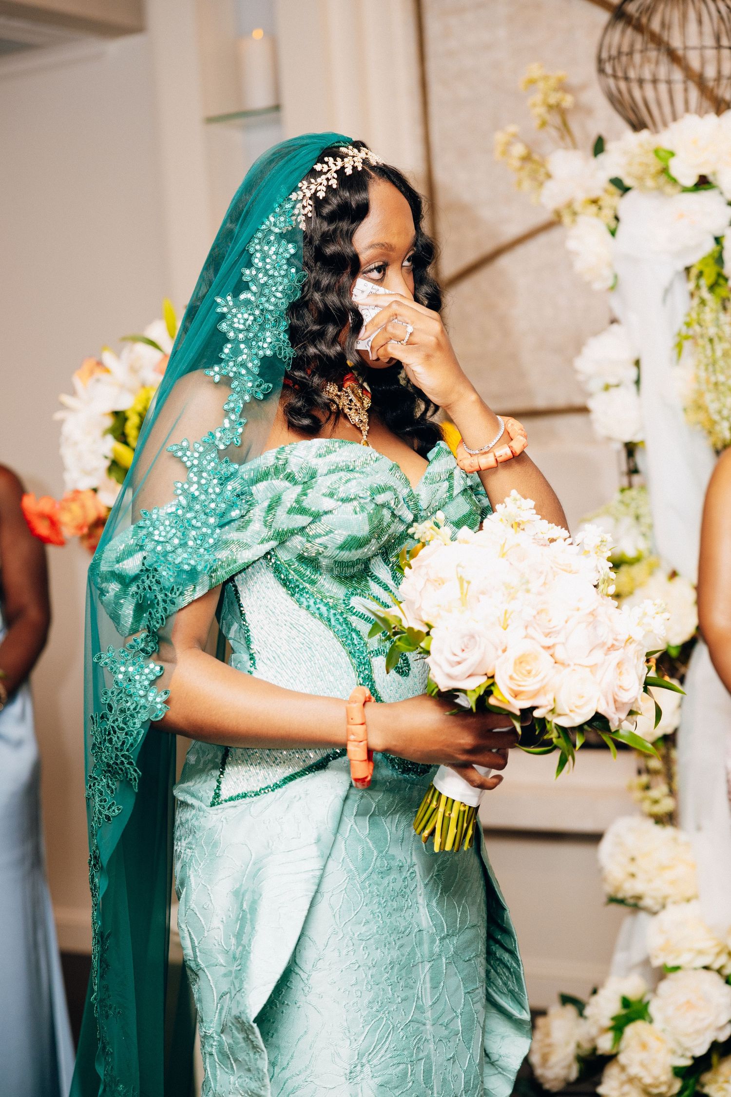 Emotional bride in teal lace gown with veil wipes tears holding white rose bouquet at wedding ceremony.