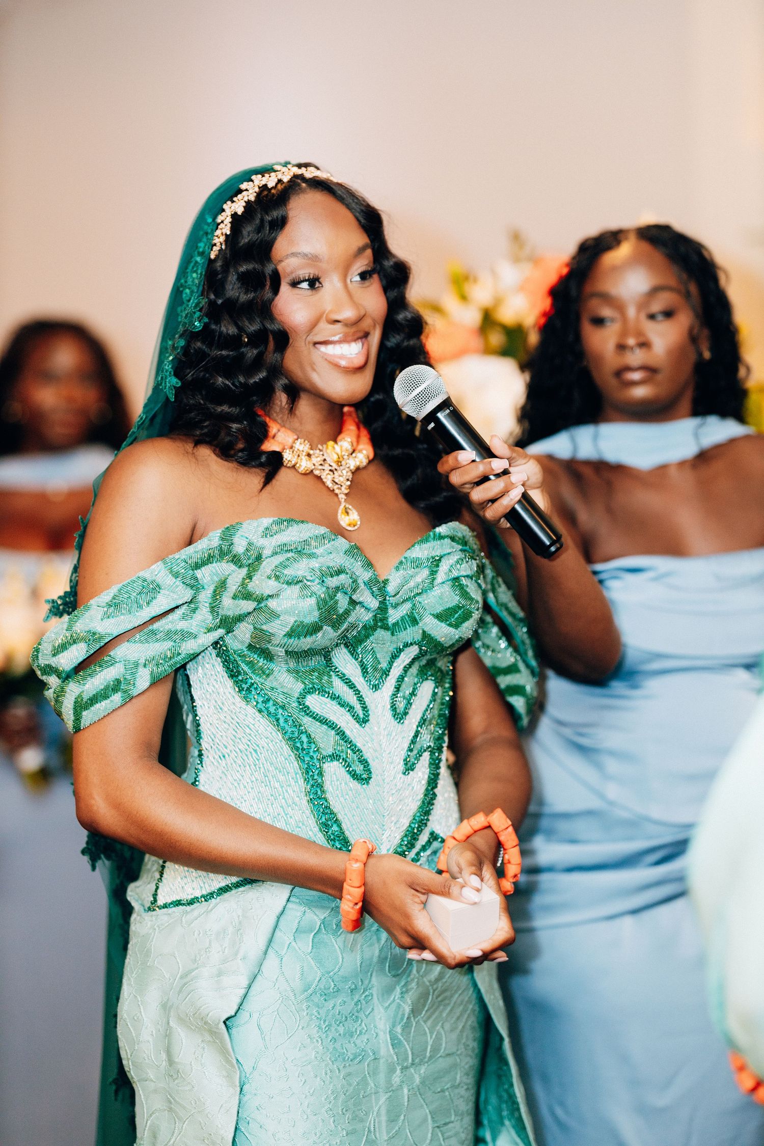Smiling bride in teal embroidered gown holds microphone at wedding with bridesmaids in blue.
