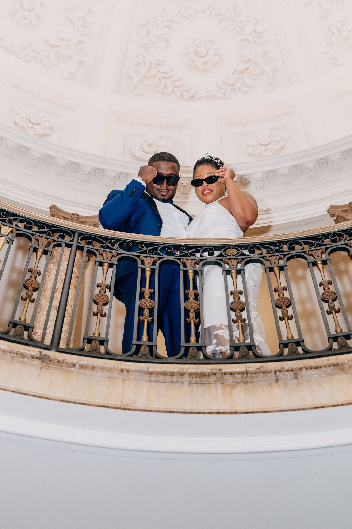 Stylish couple in wedding attire posing with sunglasses on an ornate balcony with decorative ceiling.