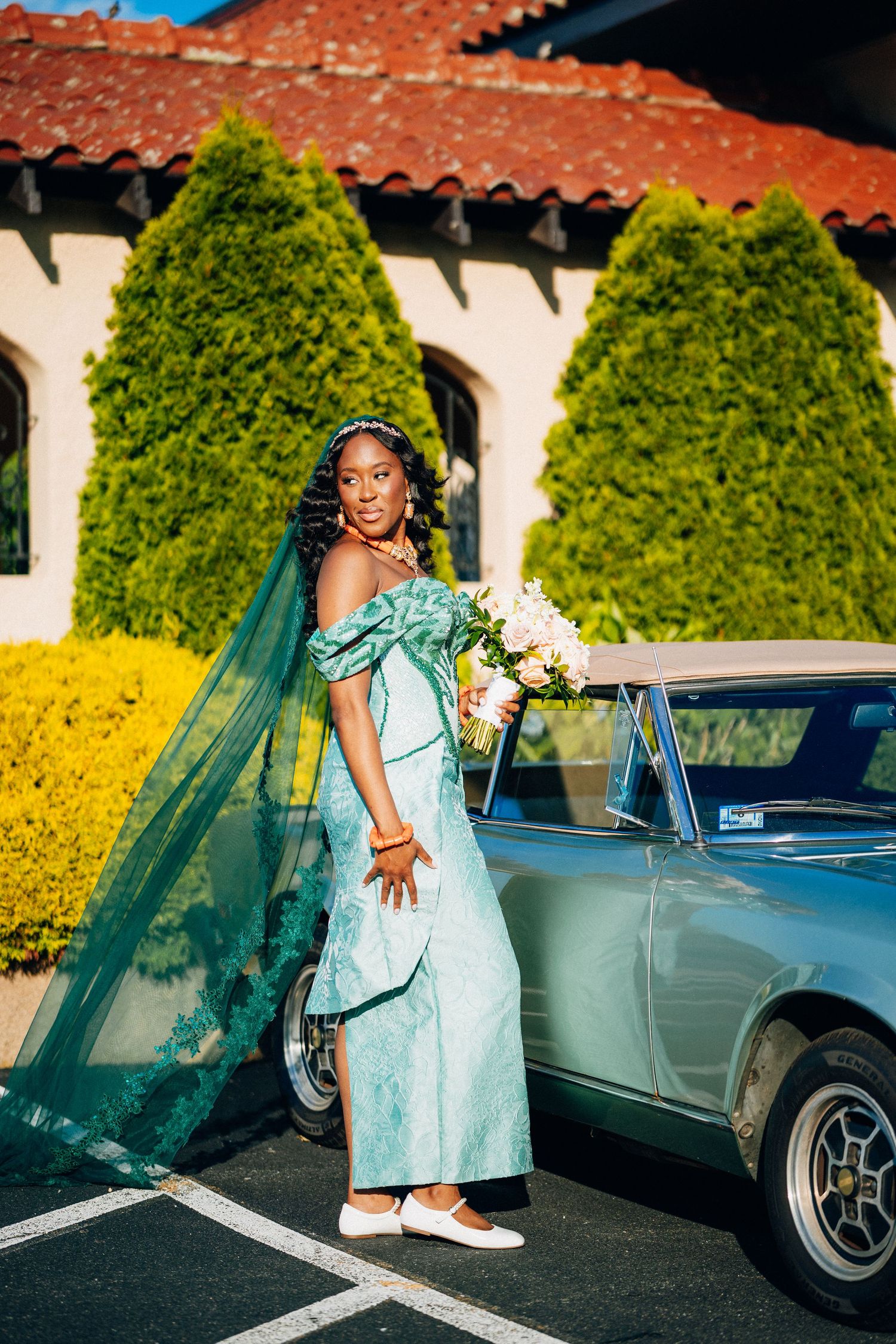 Bride in teal off-shoulder gown with flowing veil holds bouquet beside a vintage blue car at wedding venue.