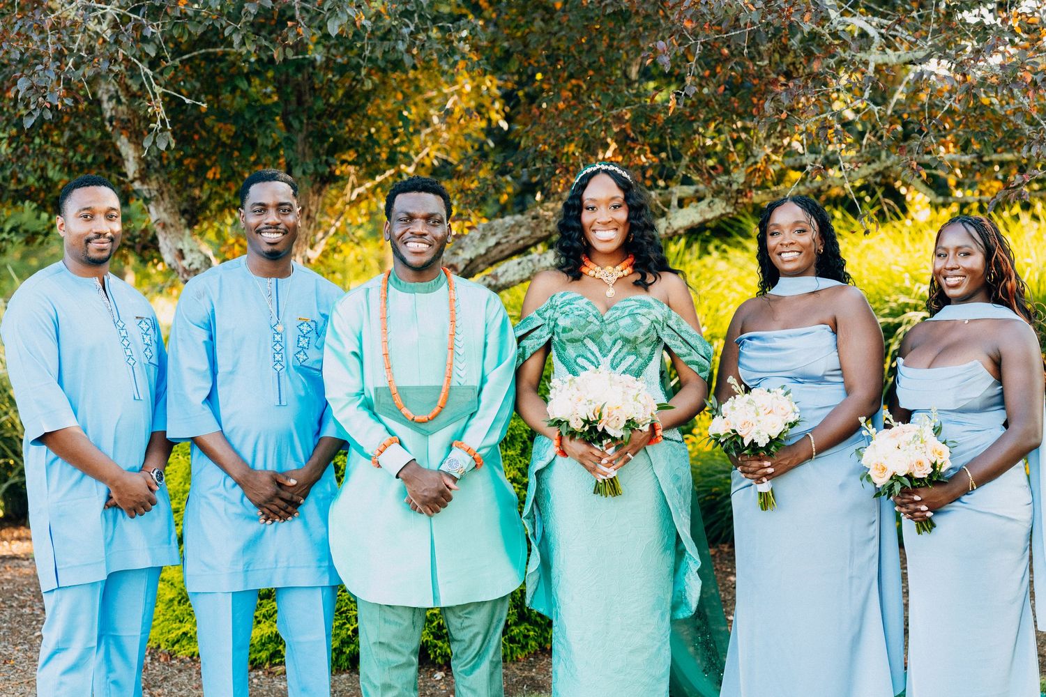 Wedding party in traditional African attire, groom in mint green, bridesmaids in blue, holding bouquets outdoors.